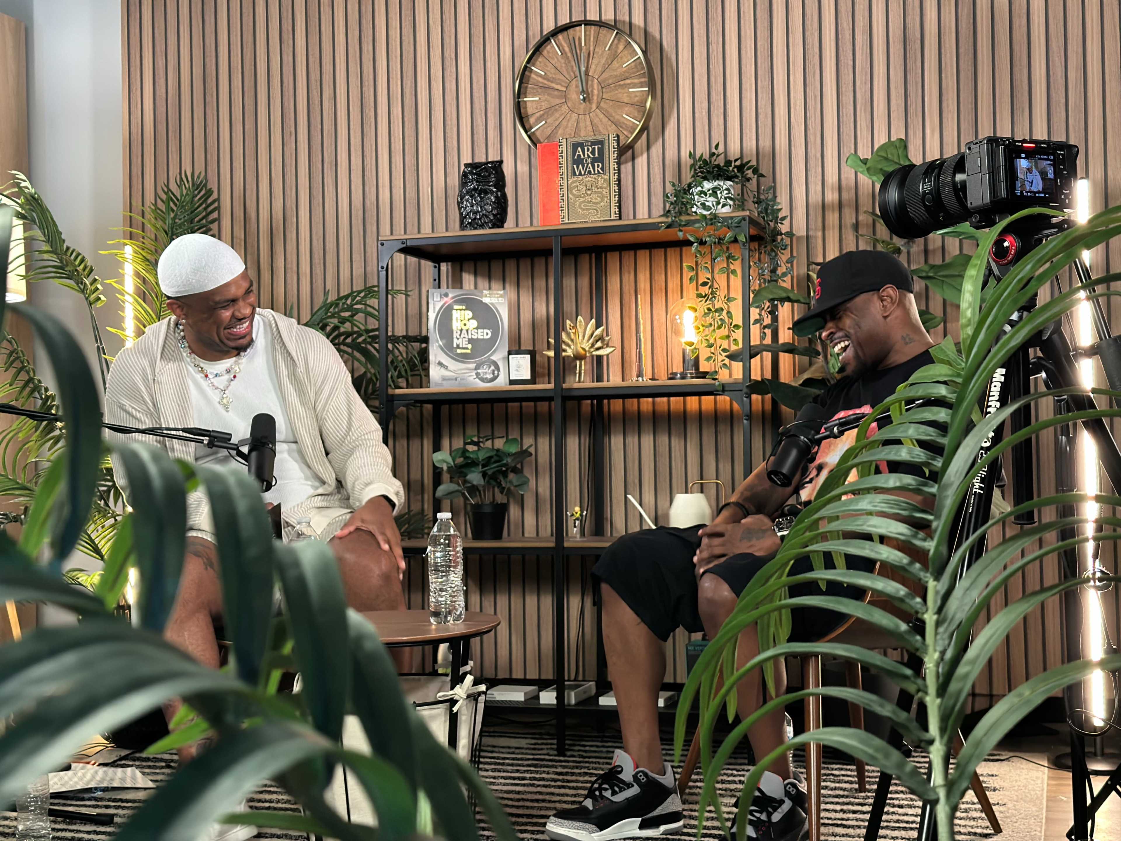 Two men are seated in a modern, indoor setting with greenery, engaged in conversation while surrounded by decorative shelves and a large camera positioned for recording.