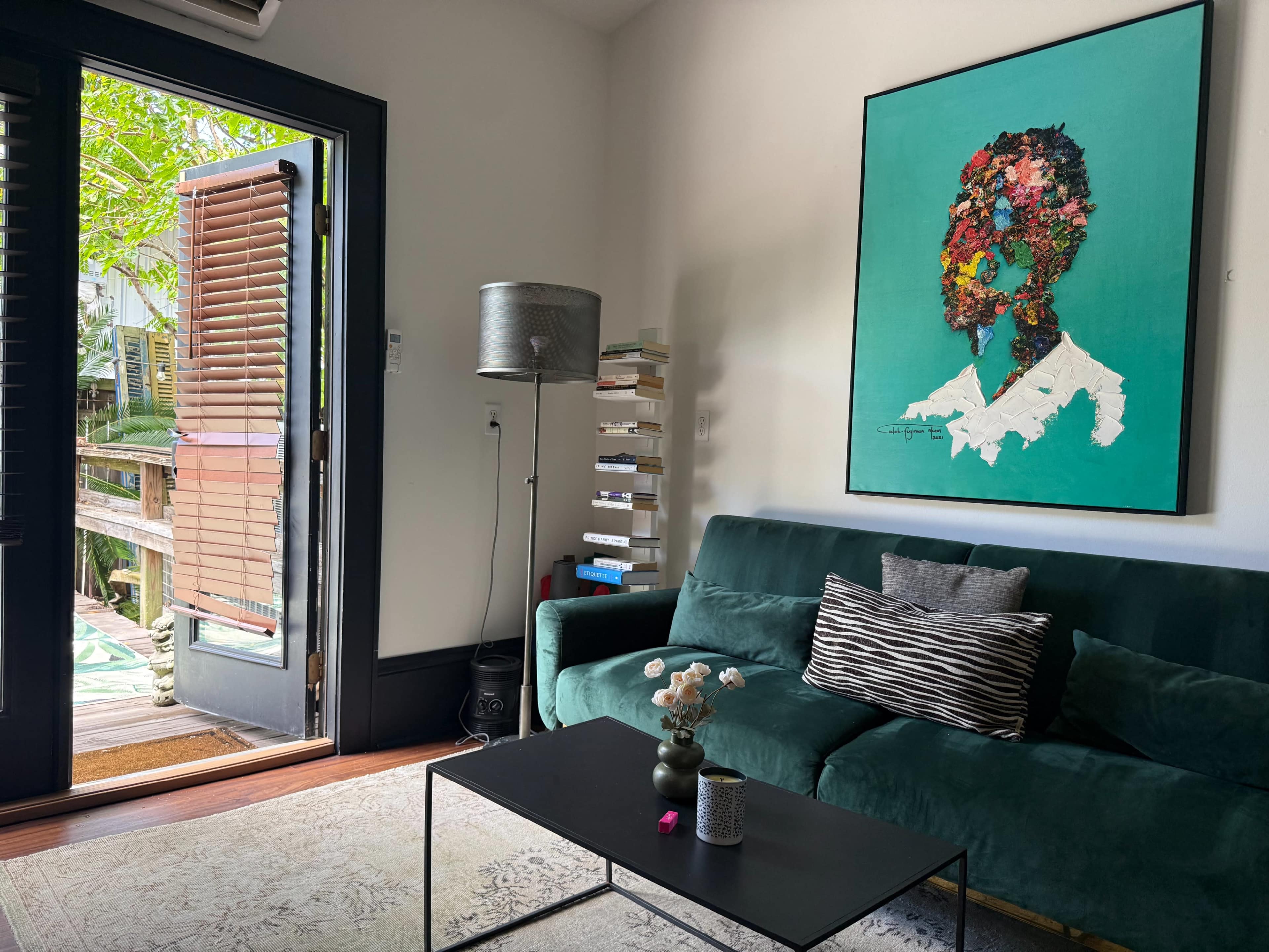 The image shows a cozy living room with a green sofa, a coffee table, a colorful abstract portrait on the wall, and a door leading to an outdoor area.