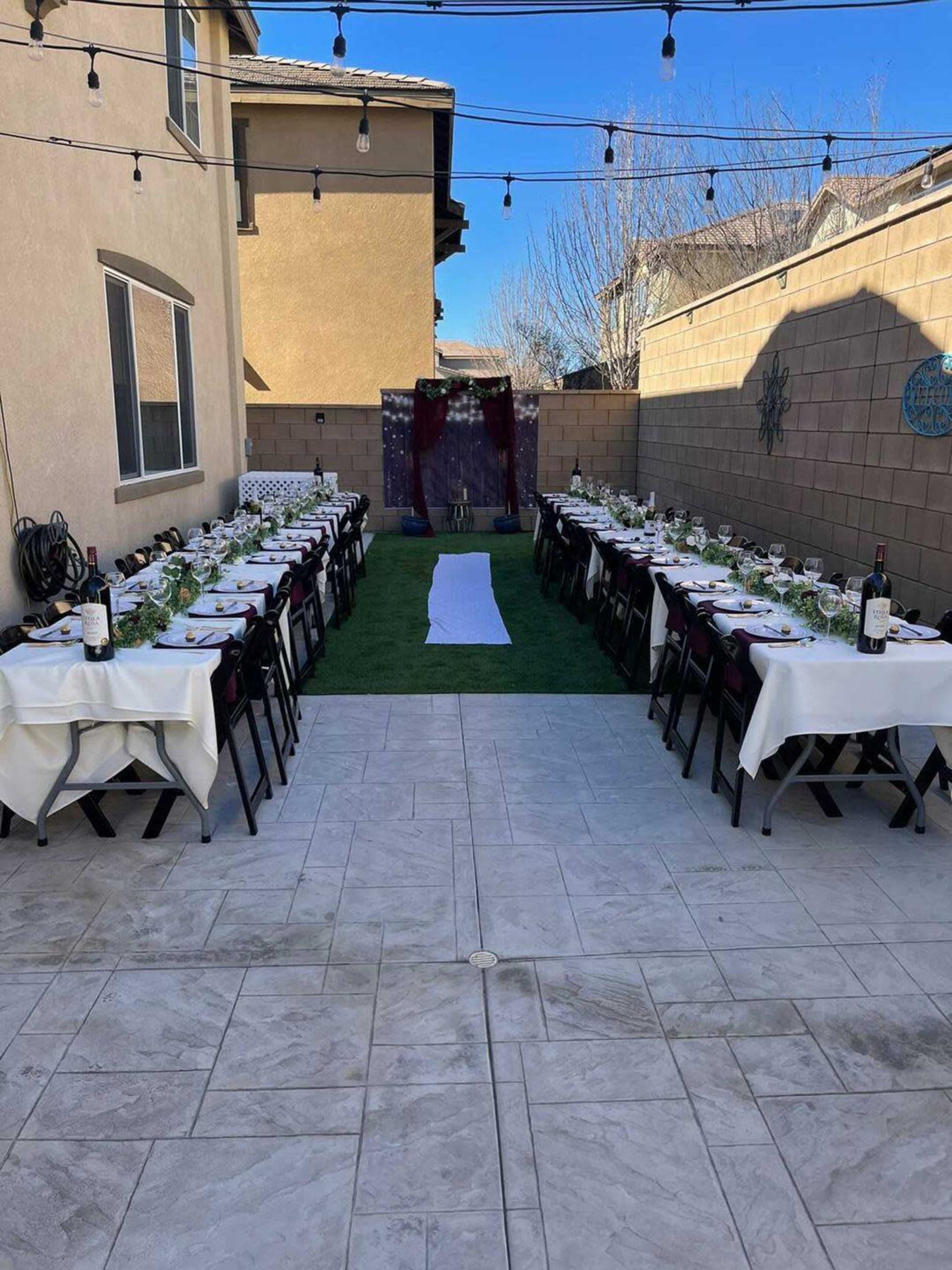 A long, elegantly set outdoor dining table is arranged on grass between two buildings, with string lights overhead and decorative elements in the background.