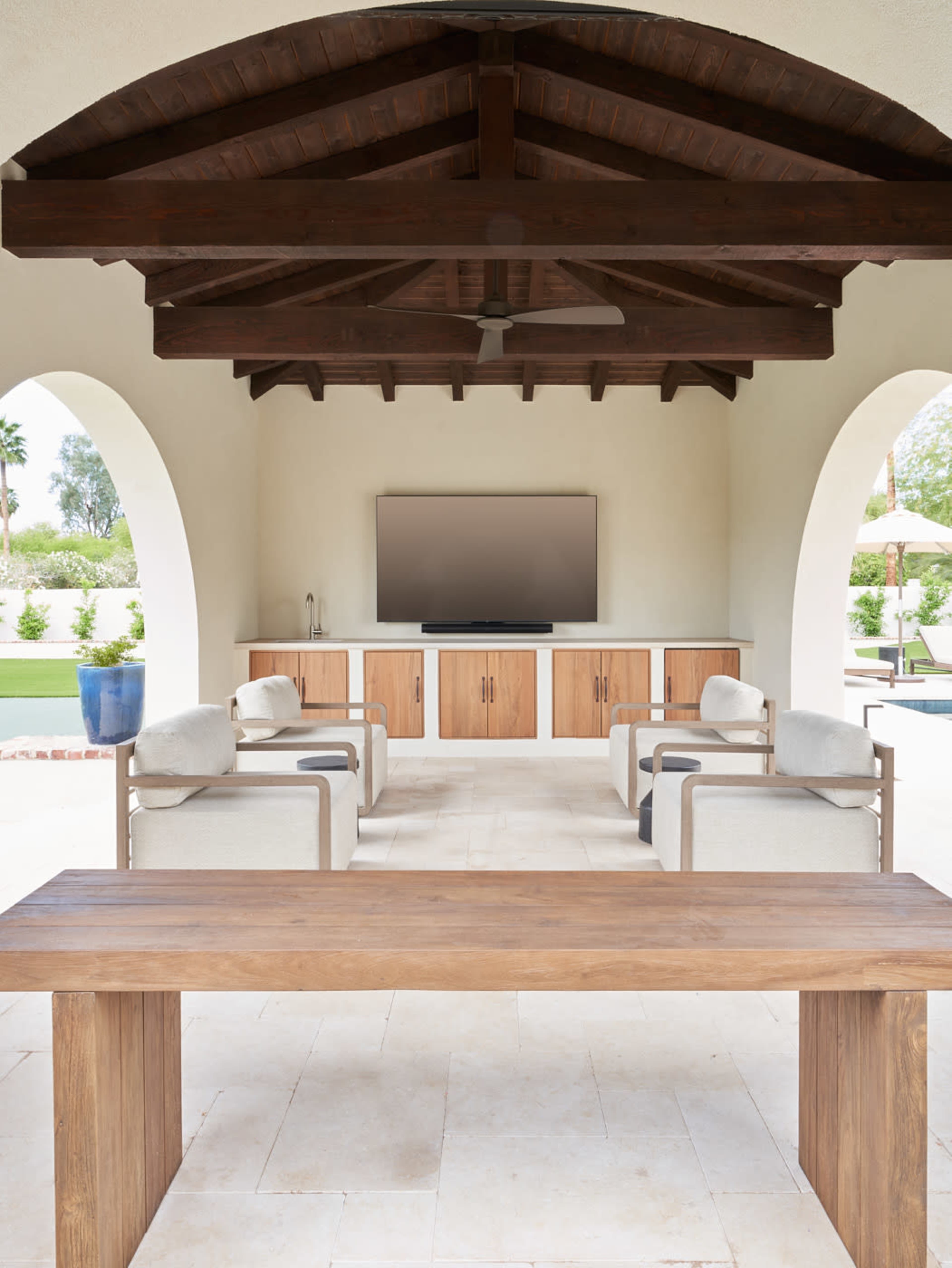 The image shows an outdoor lounge area with a wooden table, two armchairs, a television mounted on a cabinet, and arched openings leading to a landscaped garden.