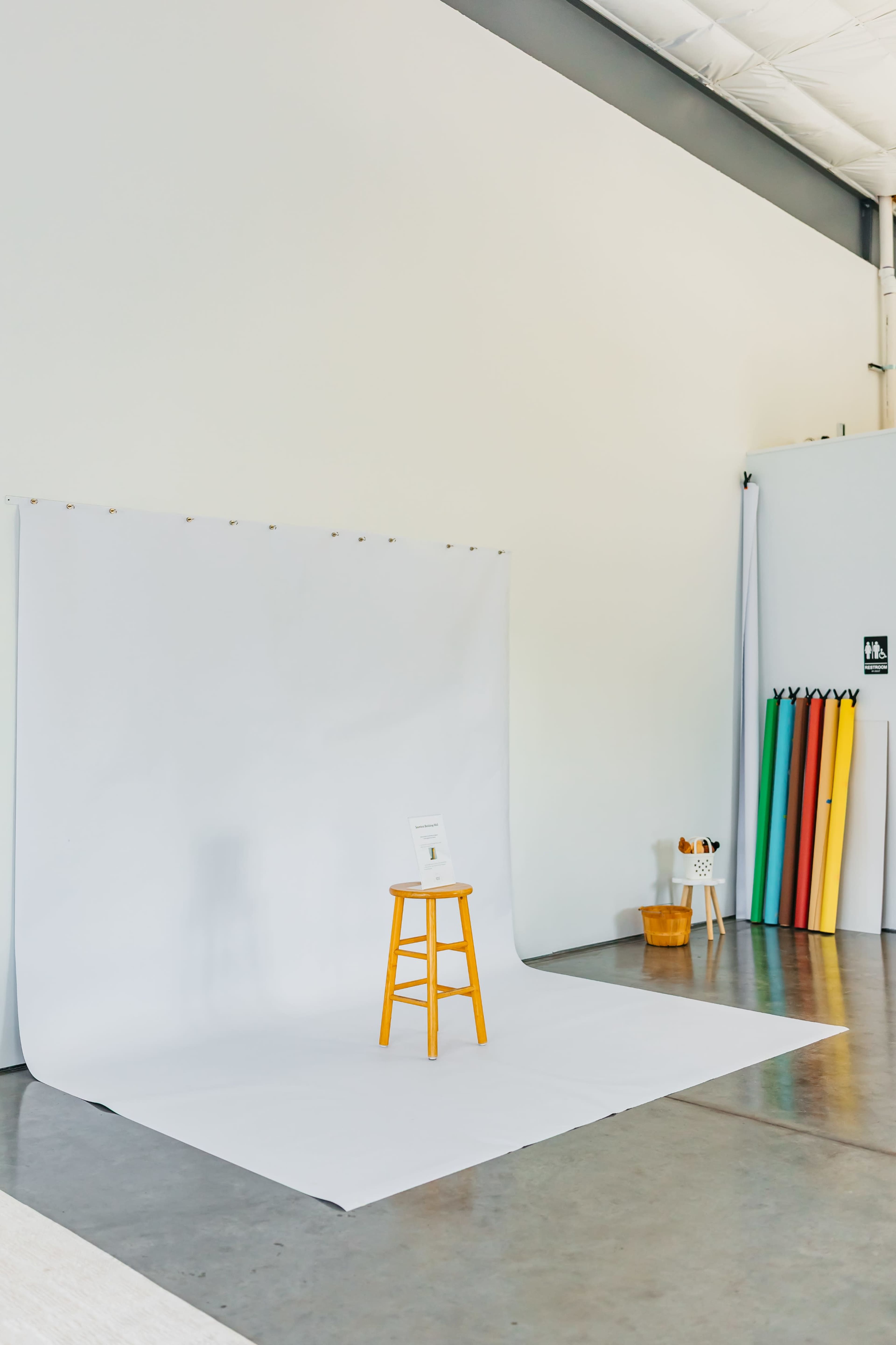 A simple studio setup features a white backdrop, a wooden stool, and colorful rolls of paper against a wall.