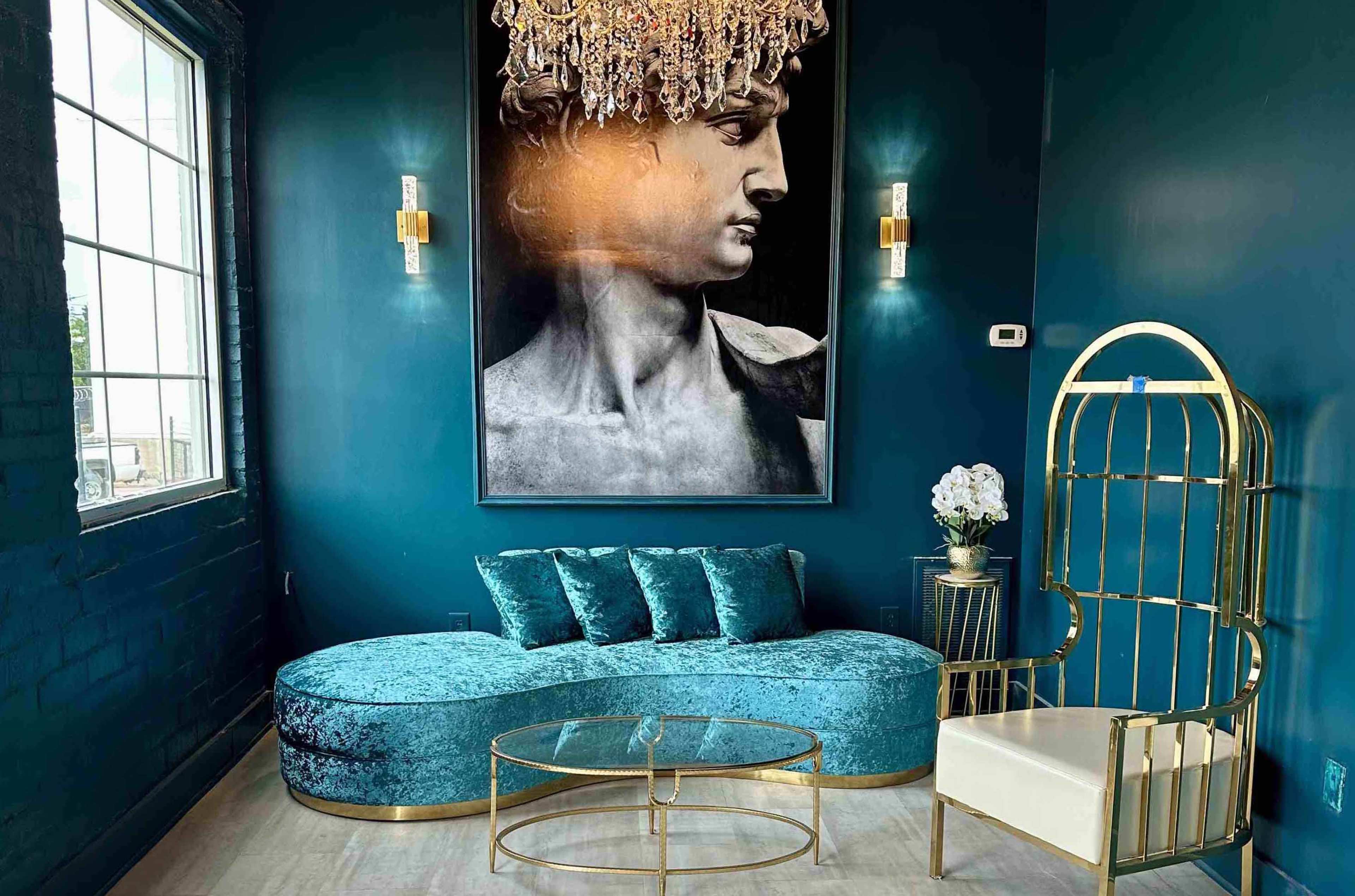 A modern lounge area featuring a teal curved sofa, a glass coffee table, a gold-framed chair, and a large black-and-white portrait of a male figure on the wall under a chandelier.