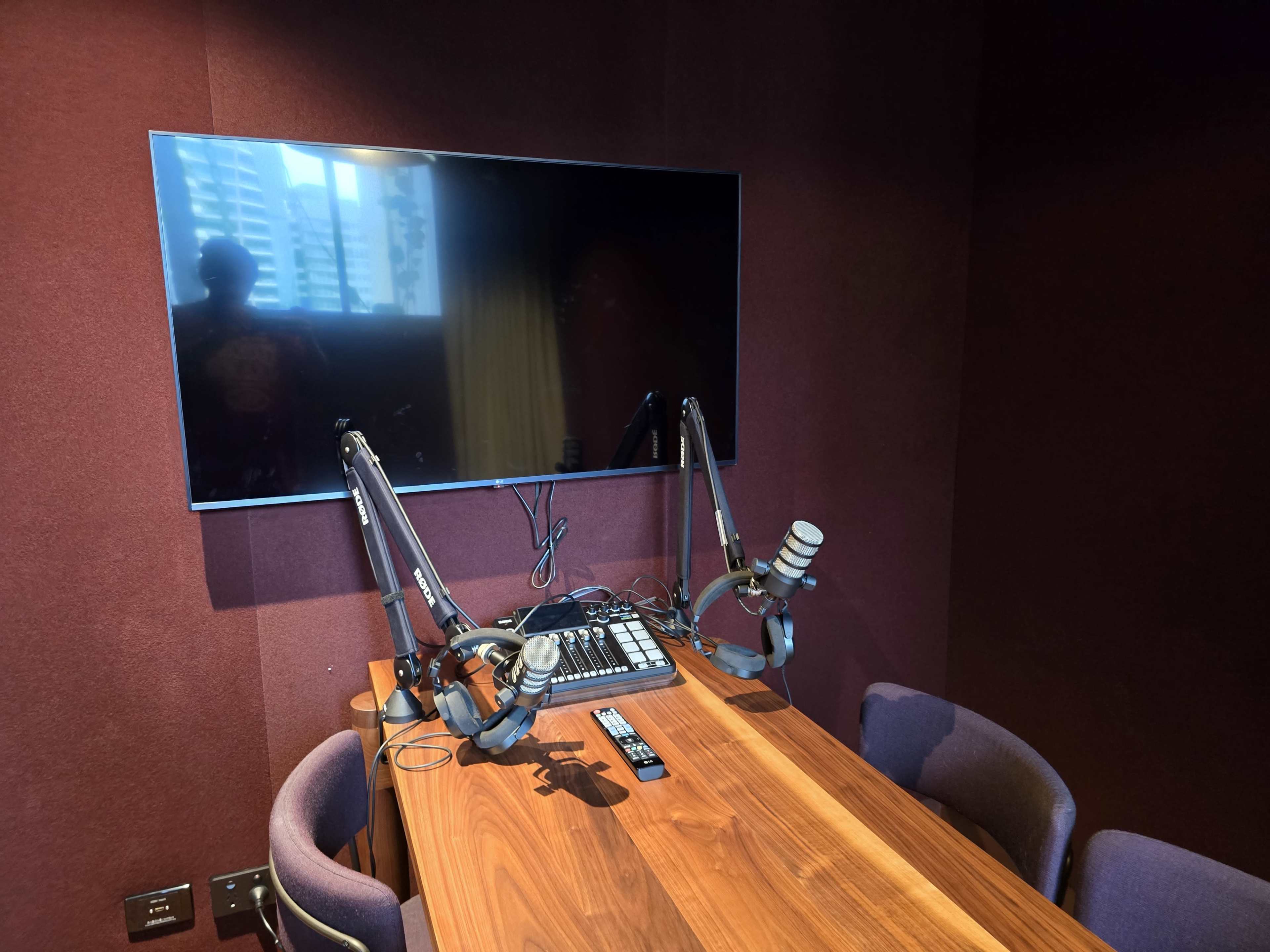 The image shows a recording studio with a wooden table, three microphones on adjustable arms, a mixer, and a large flat-screen TV mounted on the wall.
