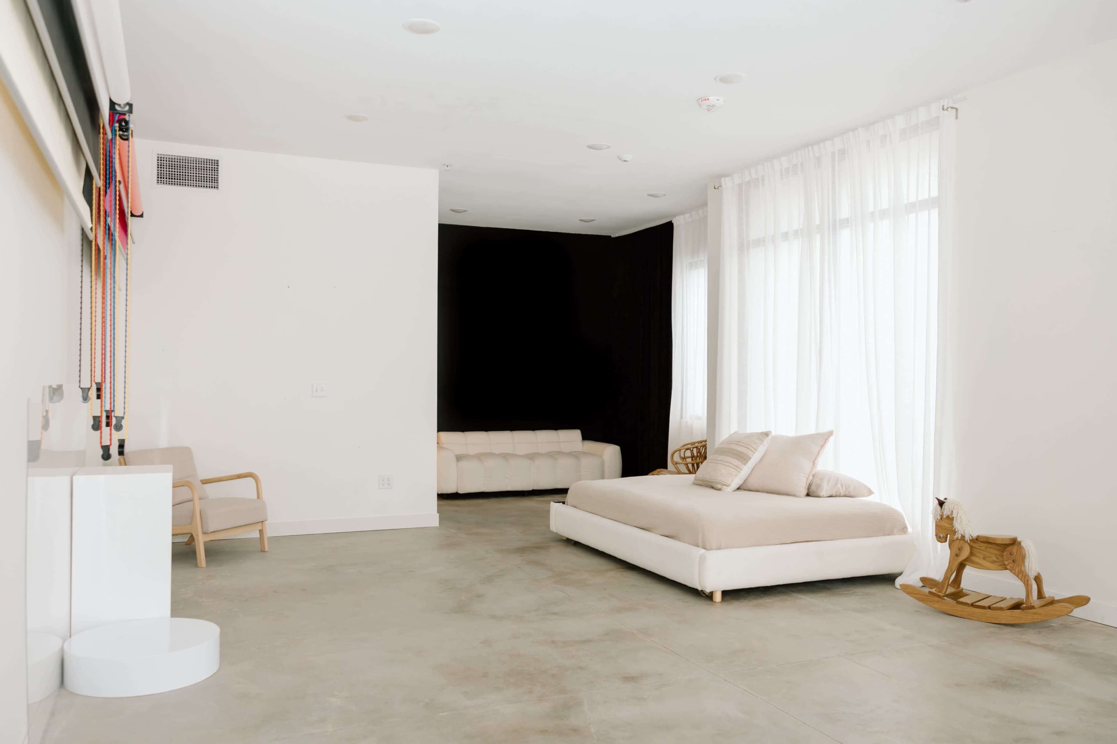 The image shows a minimalistic bedroom featuring a bed with a light-colored mattress, a small couch in the background, large windows with sheer curtains, and a wooden rocking horse on the floor.