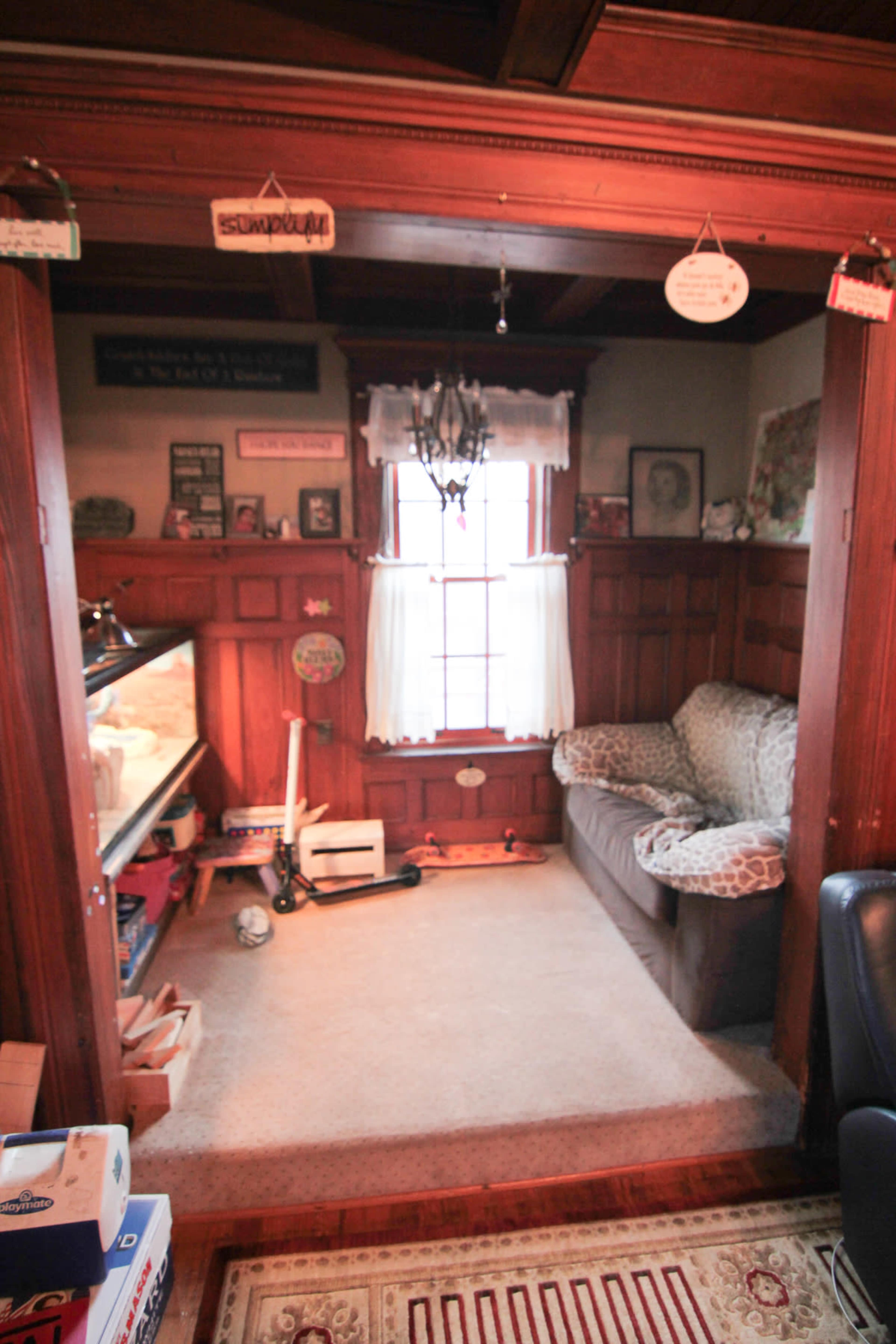 A small, cozy room featuring wooden paneling, a sofa, and a window with sheer curtains, along with various decorations on the walls.