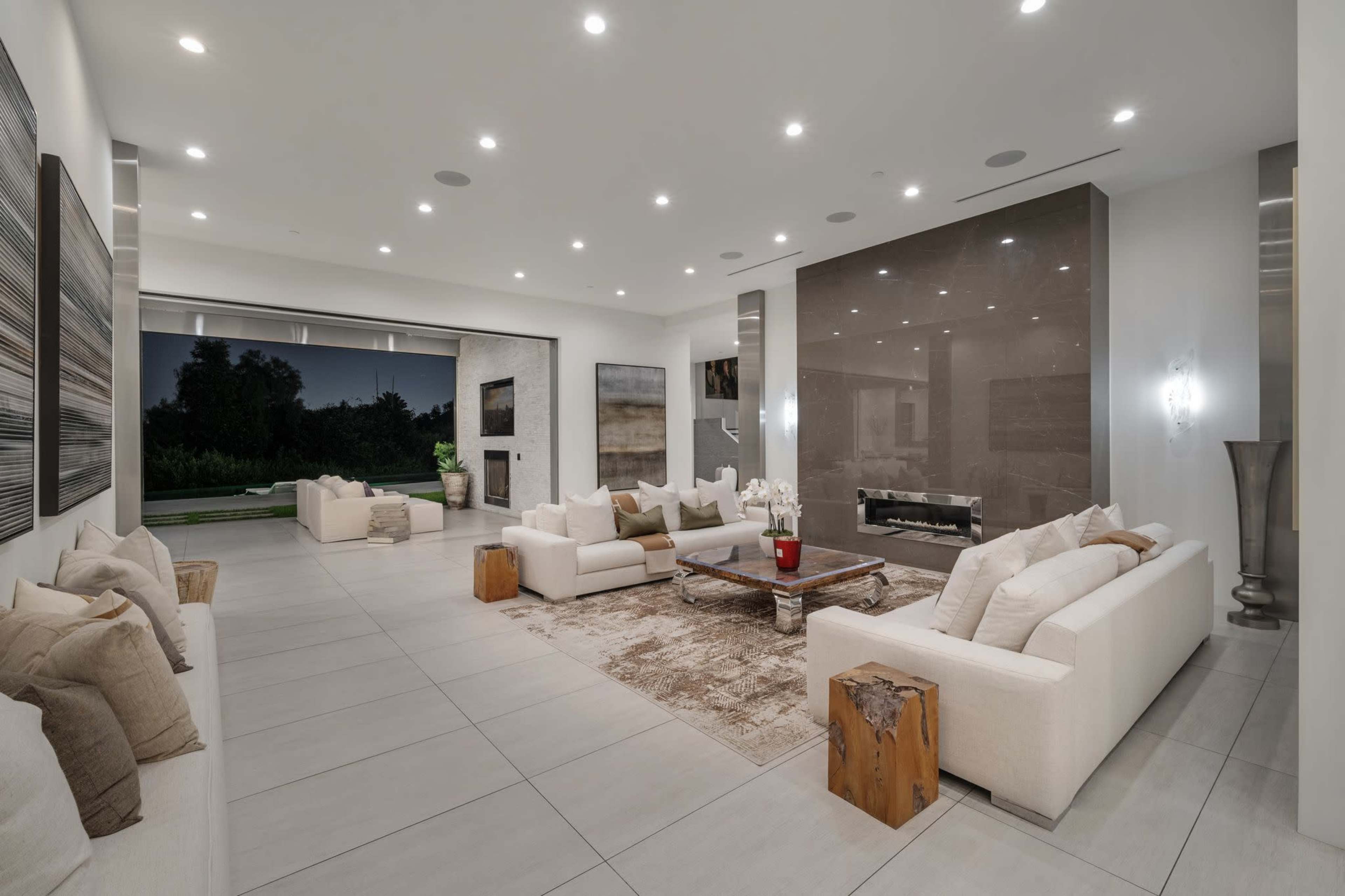 The image shows a modern living room featuring white sofas, a central wooden coffee table, a stone fireplace, and large windows overlooking a landscaped area.