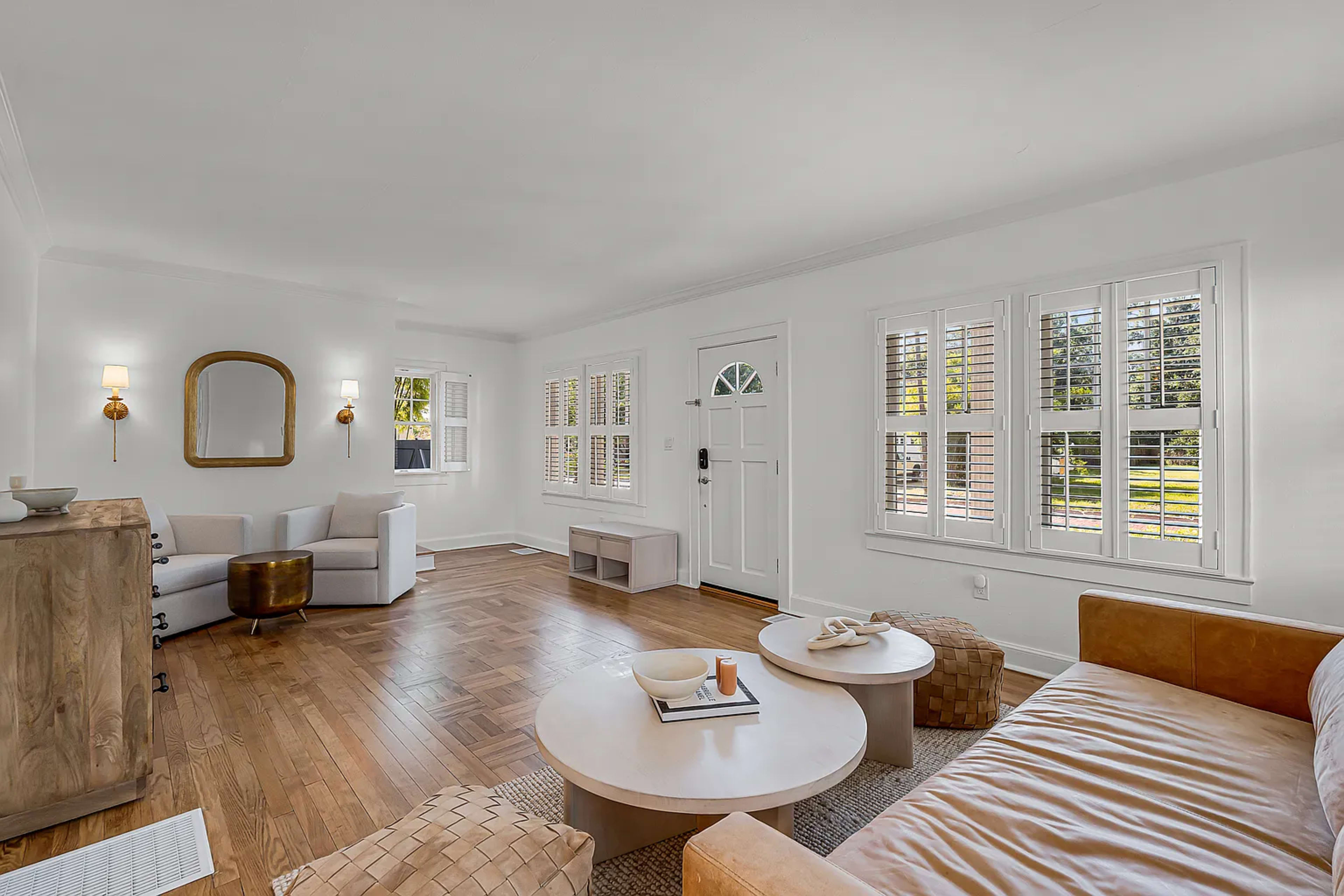 The image shows a well-lit living room featuring a tan leather sofa, a round coffee table, and light-colored walls with window shutters.