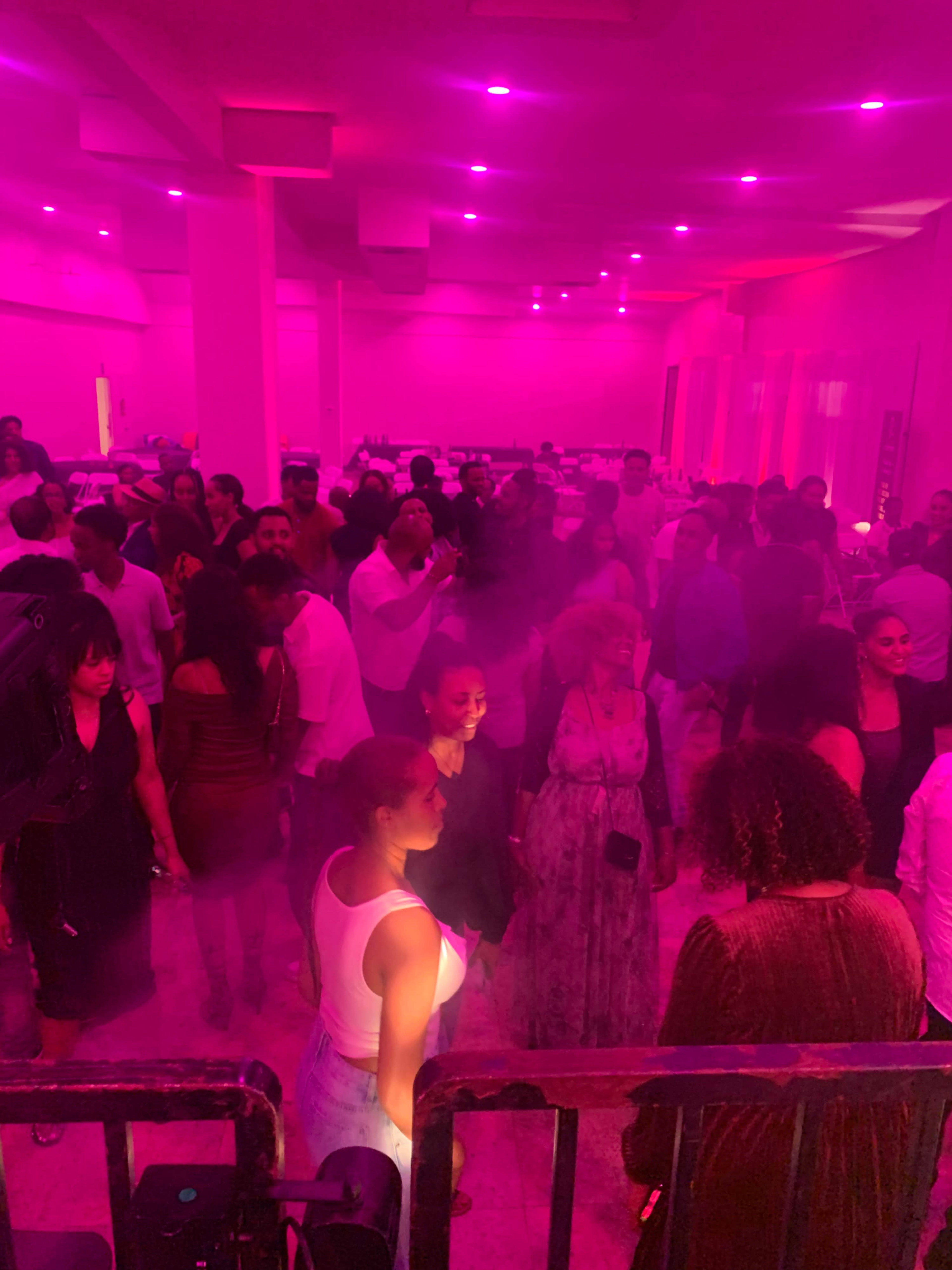A crowded venue filled with people dancing under pink lighting and fog effects.
