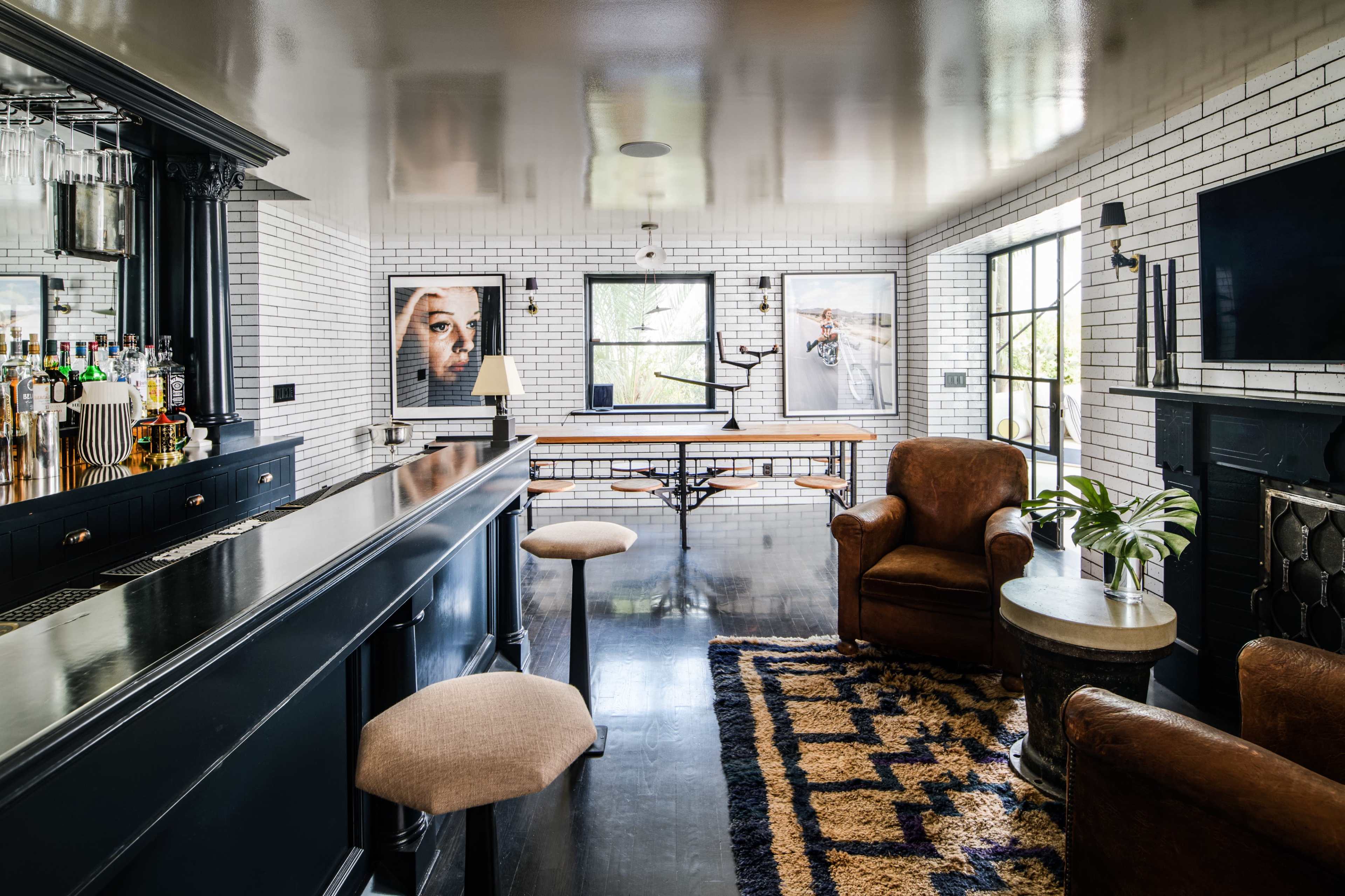 The image features a modern bar area with a black counter, two upholstered stools, and a seating arrangement that includes a brown armchair, situated in a room with white tiled walls and framed artwork.