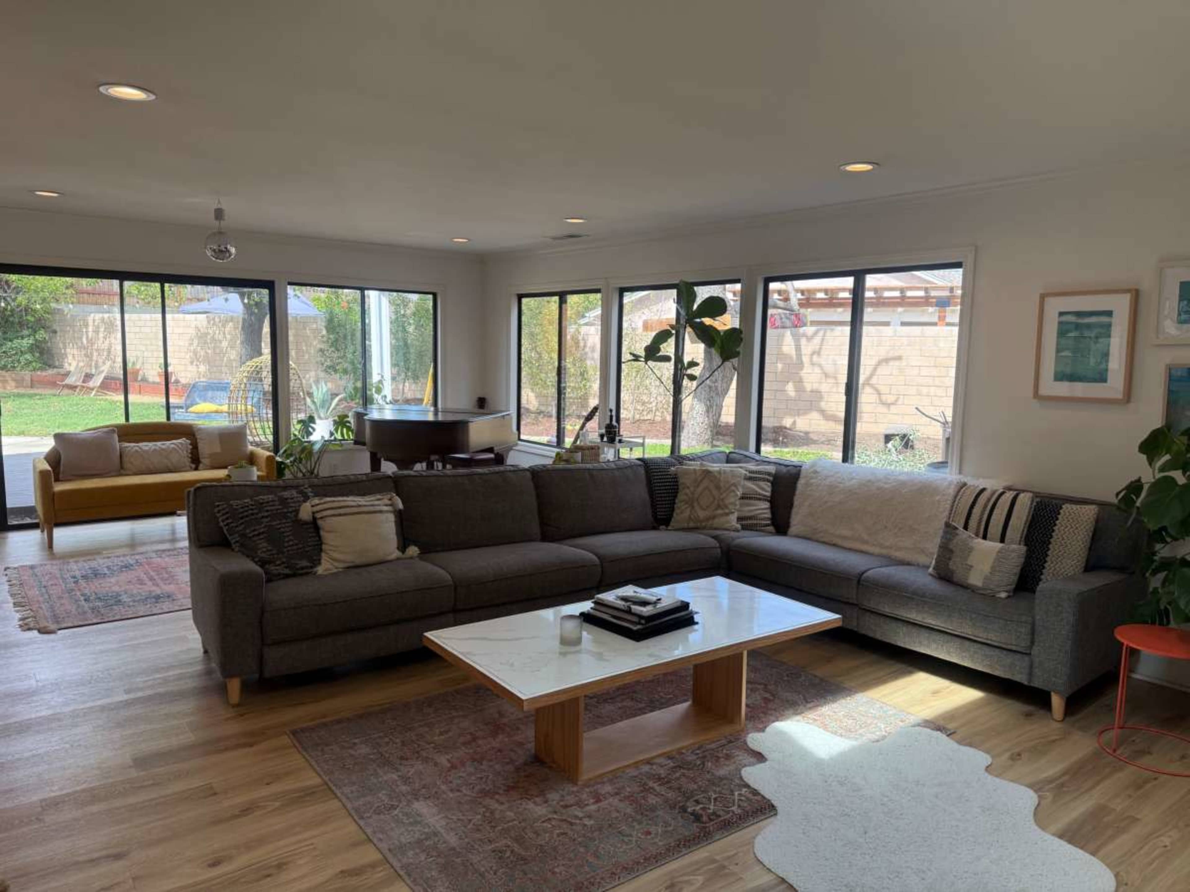 The image shows a modern living room featuring a large sectional sofa, a coffee table, and large windows that offer a view of an outdoor area with greenery.