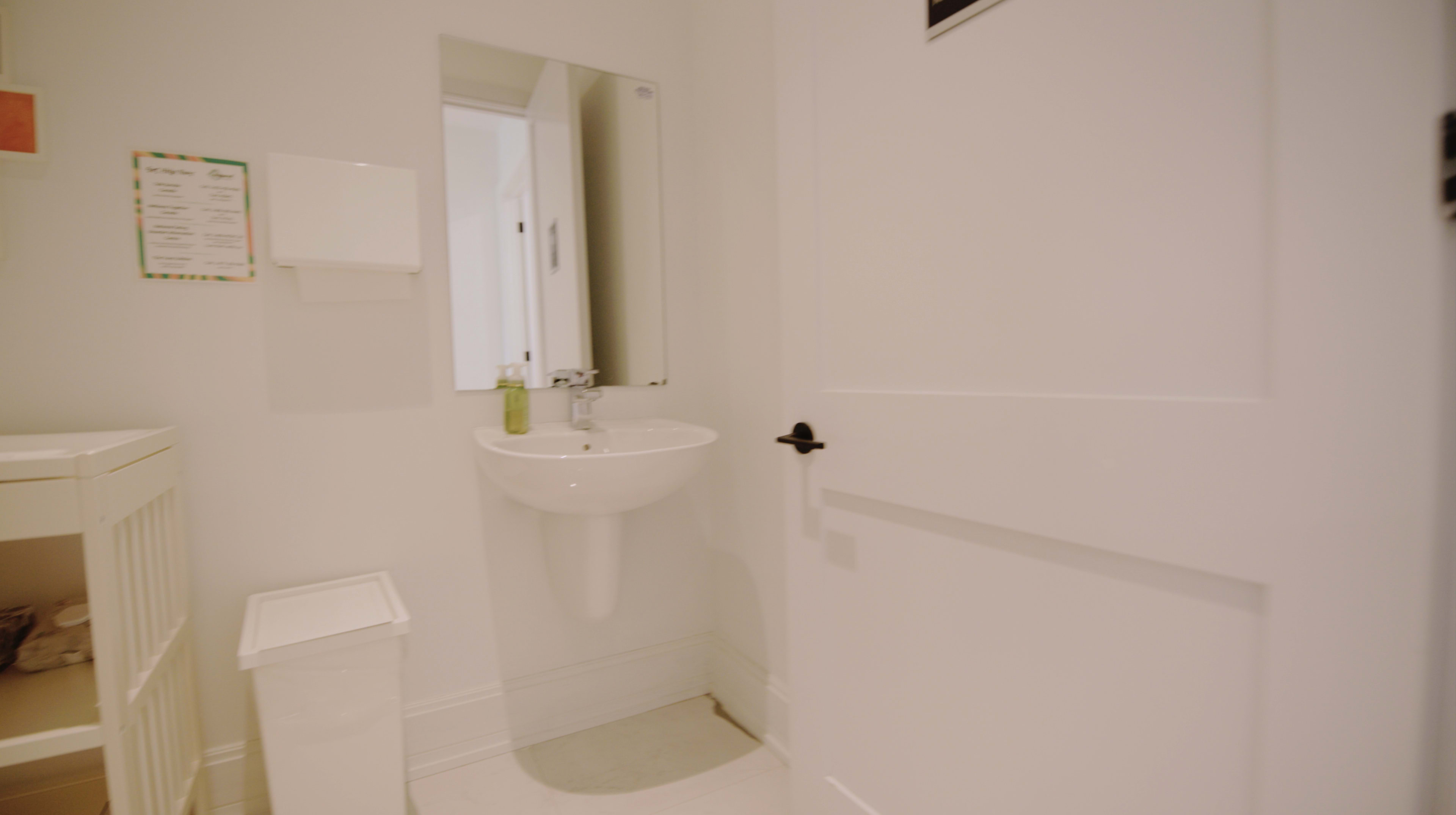 The image shows a small, minimalist bathroom with a white sink, a mirror above it, and a waste bin nearby.