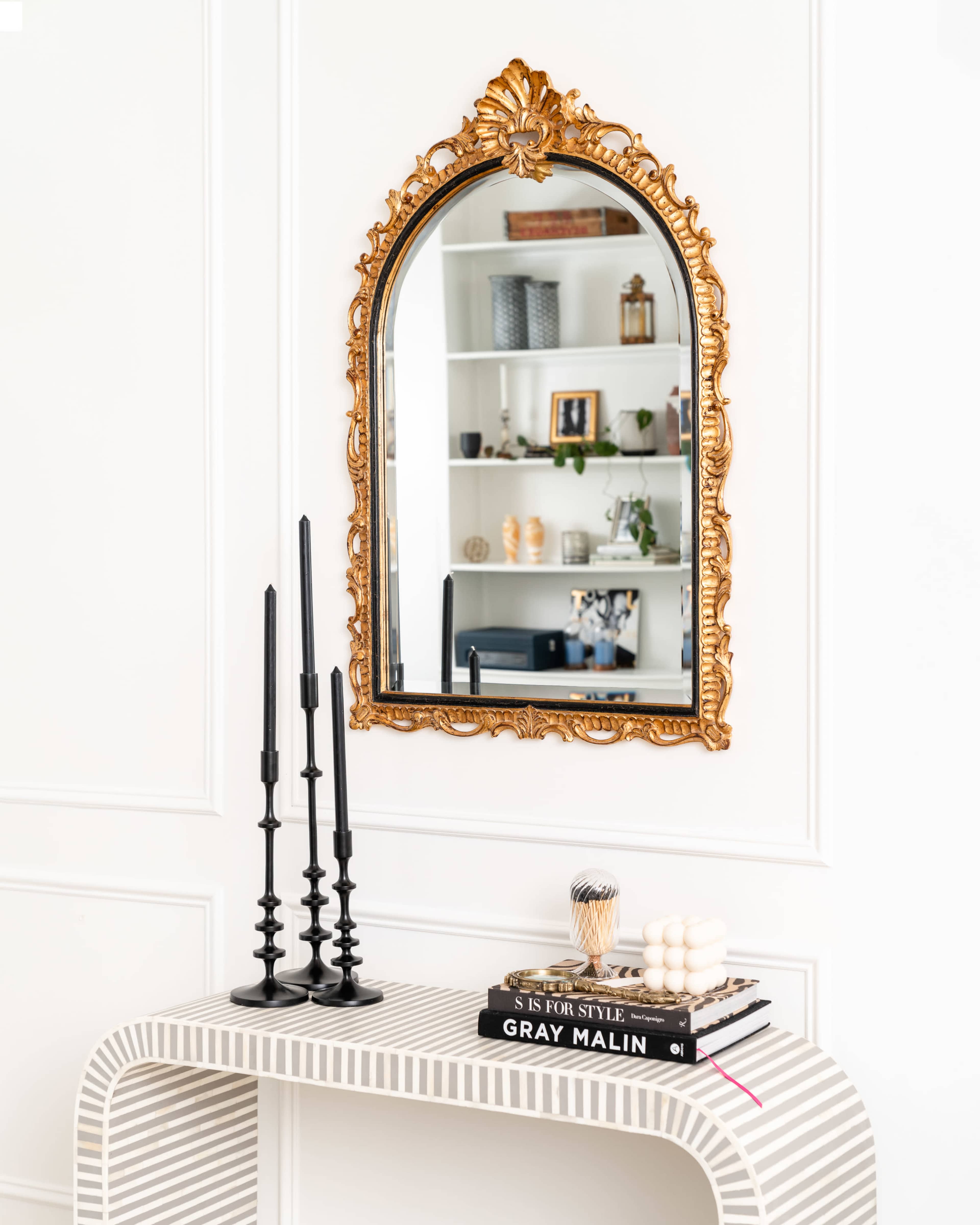 A gold-framed mirror hangs above a striped console table adorned with black candlesticks and stacked books.