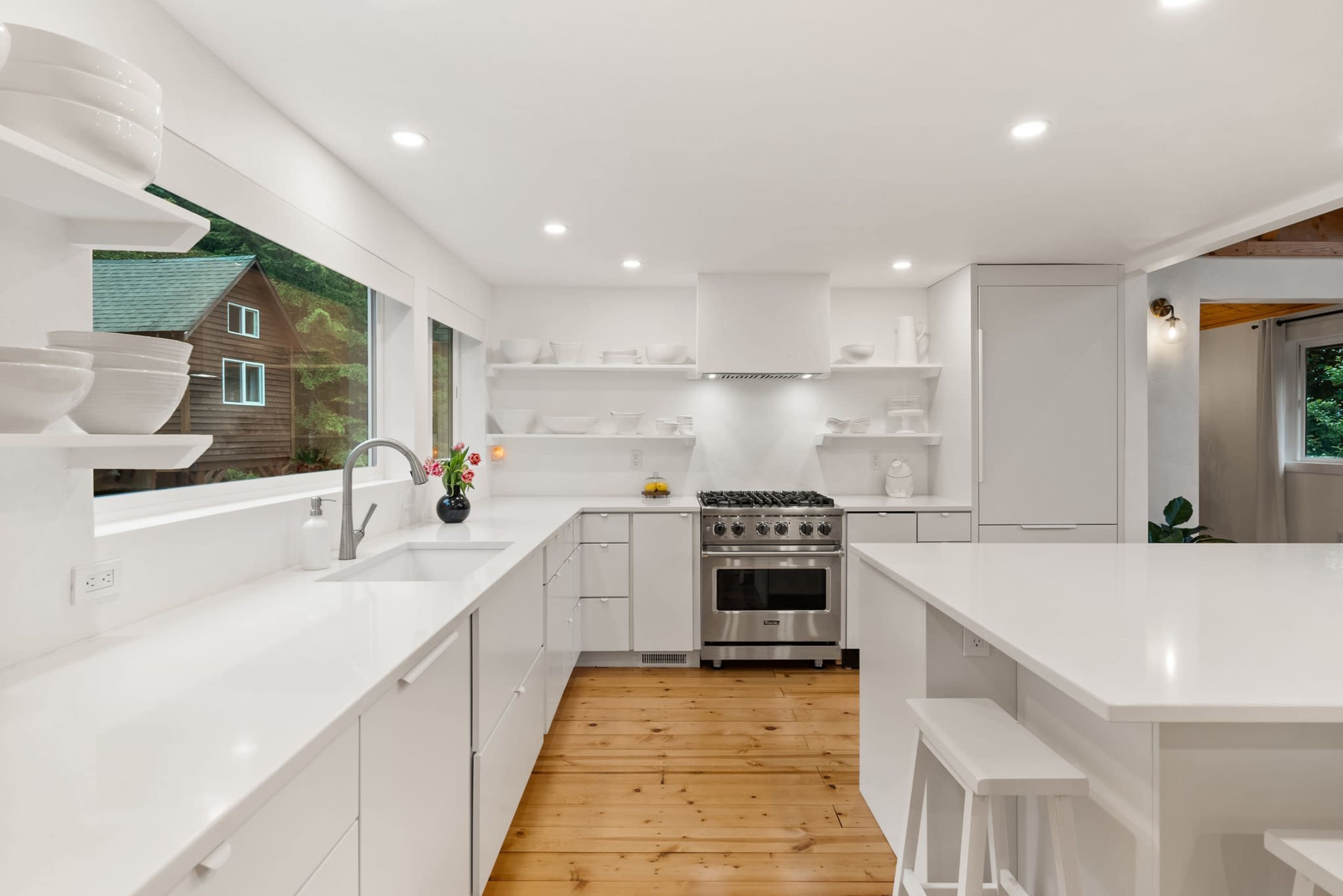 The kitchen features white cabinetry, a stainless steel stove, and open shelving, with a wood floor and large windows allowing natural light.