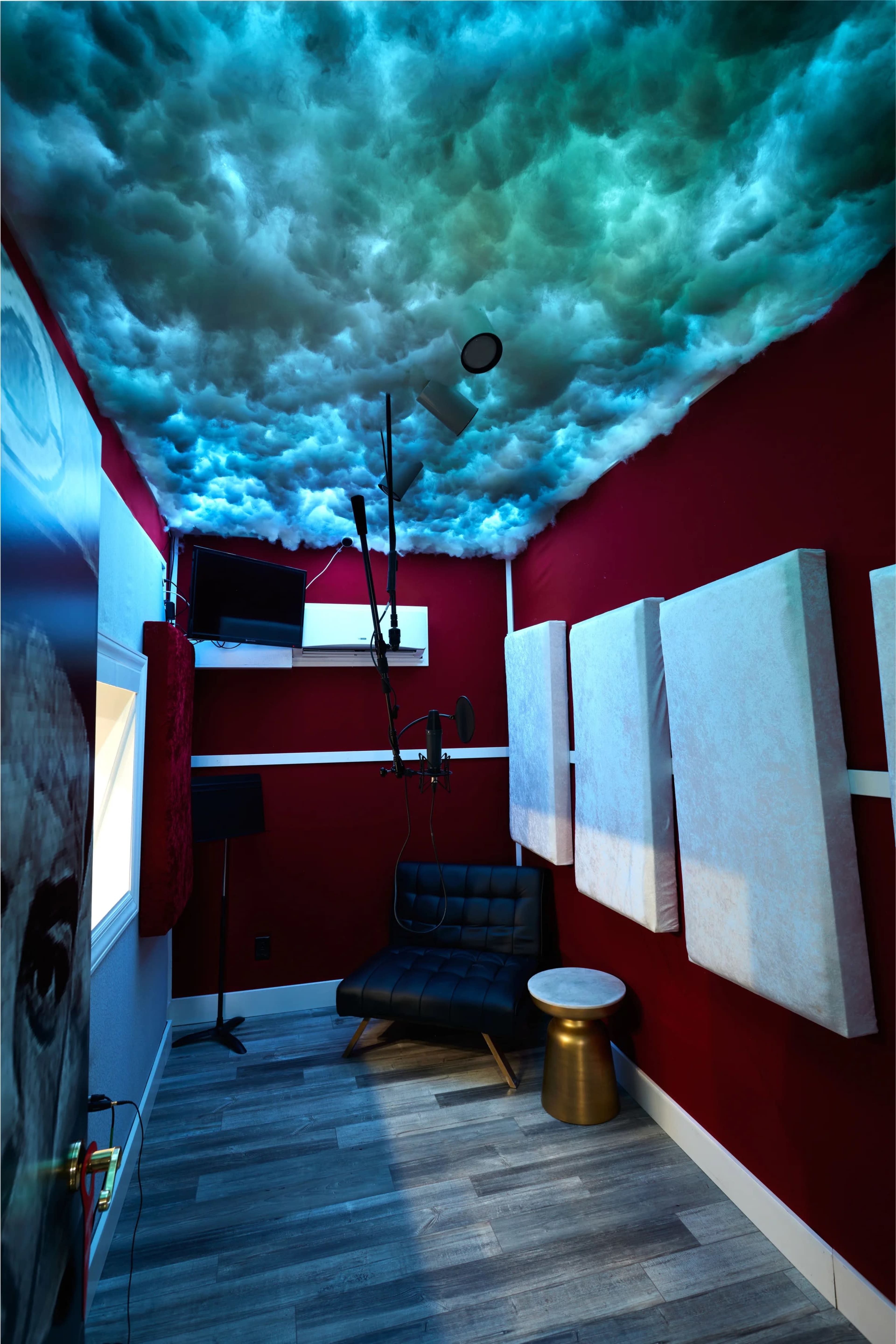 The image shows a small recording booth with a cloud-patterned ceiling, red walls, acoustic panels, a microphone setup, and a black chair beside a metal table.