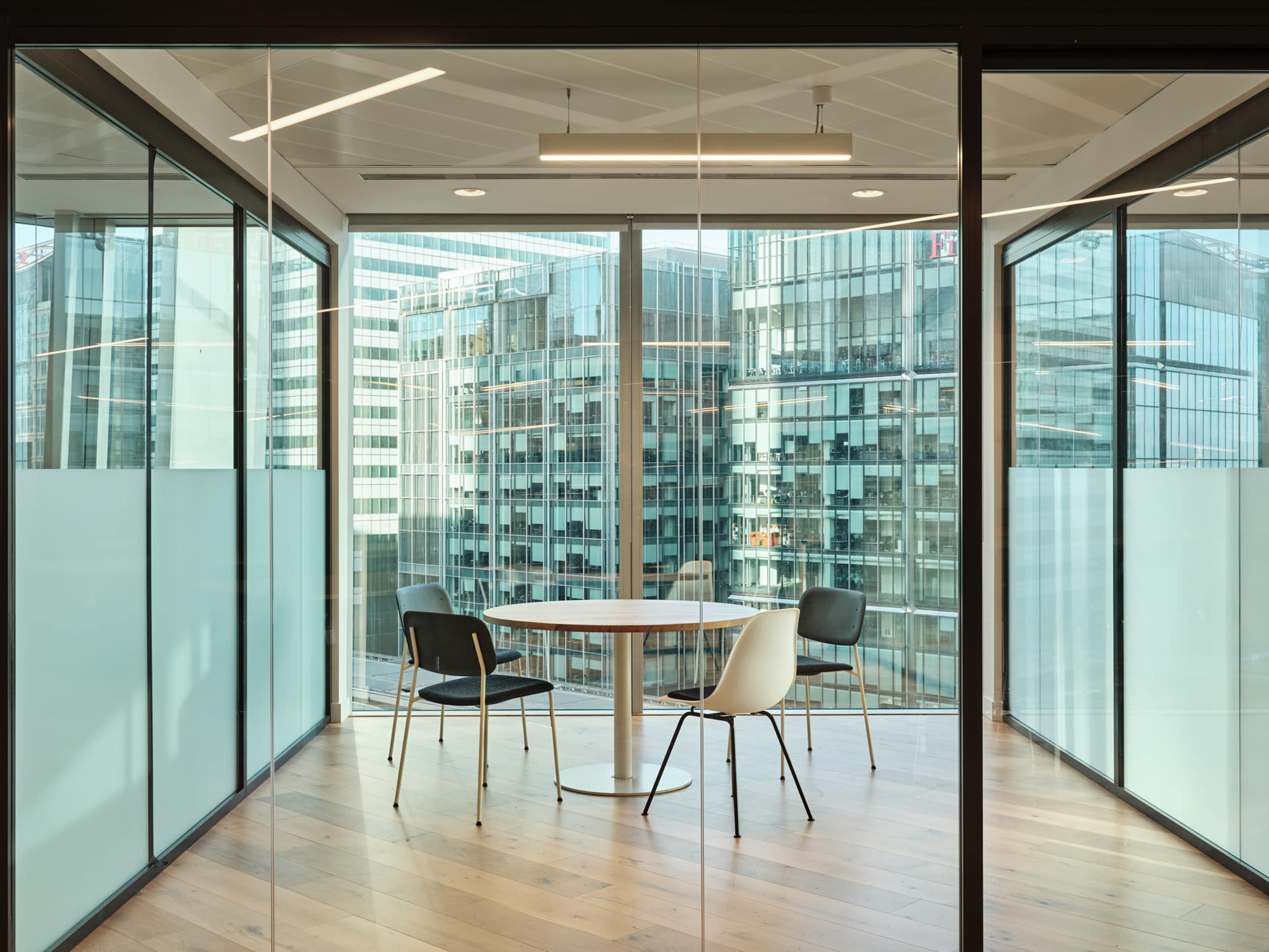 A modern meeting room with a circular table and three chairs, surrounded by large glass windows overlooking a cityscape.