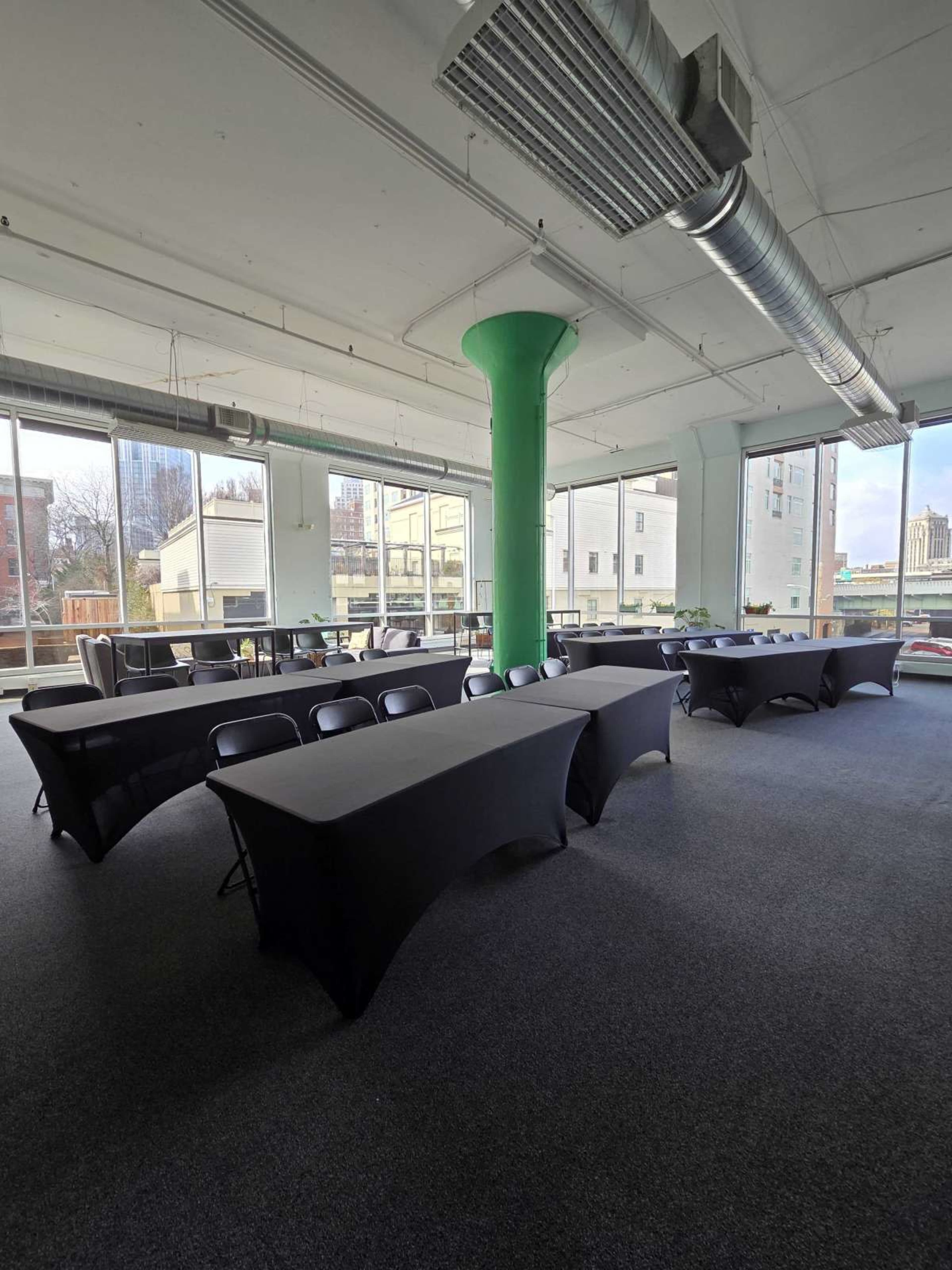 The image shows a spacious meeting room with several tables covered in black cloth, arranged in a layout suitable for a conference or gathering, along with large windows allowing natural light to enter.