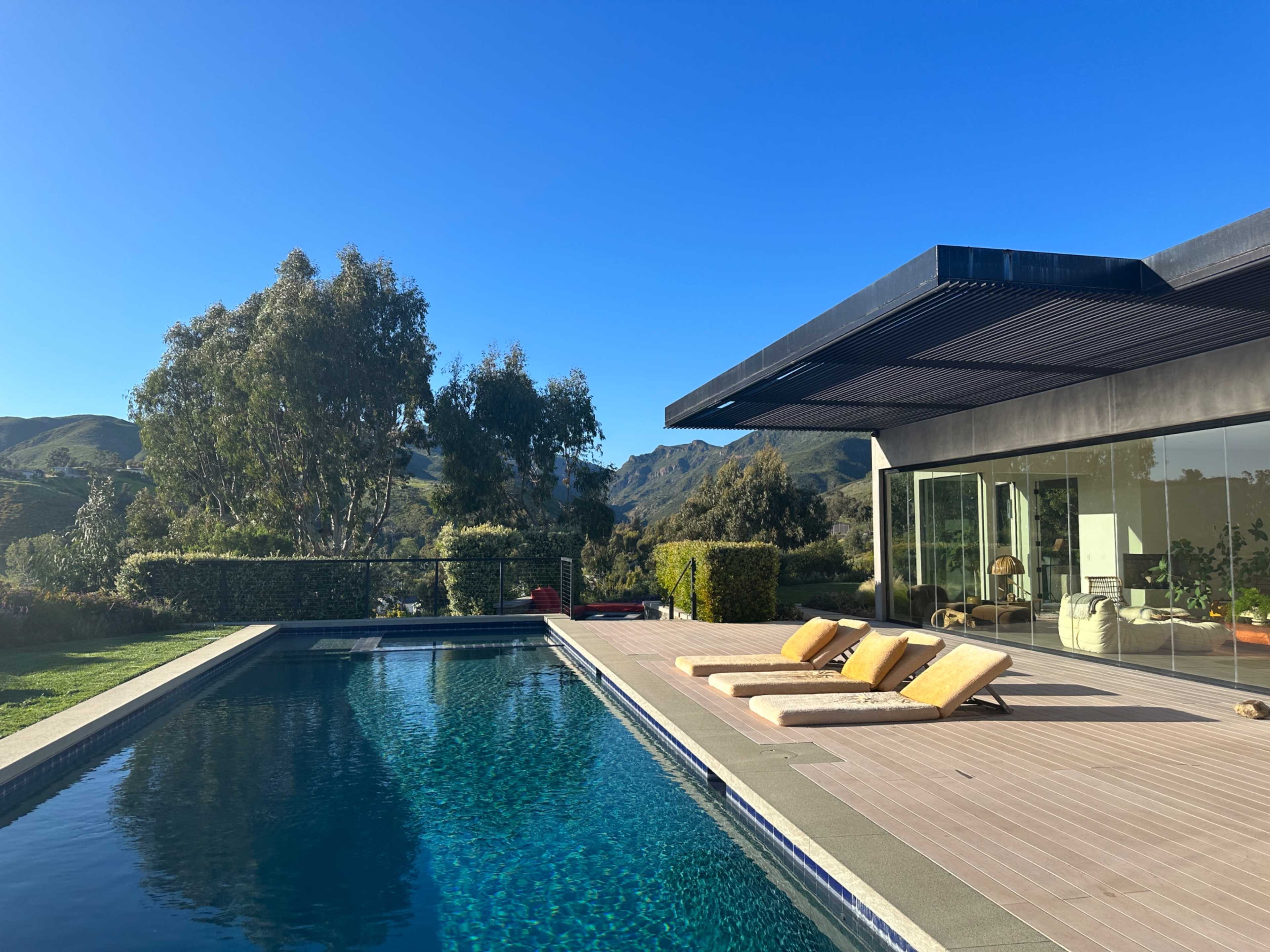 The image shows a modern house with a swimming pool, poolside loungers, and a scenic view of mountains and clear blue sky.