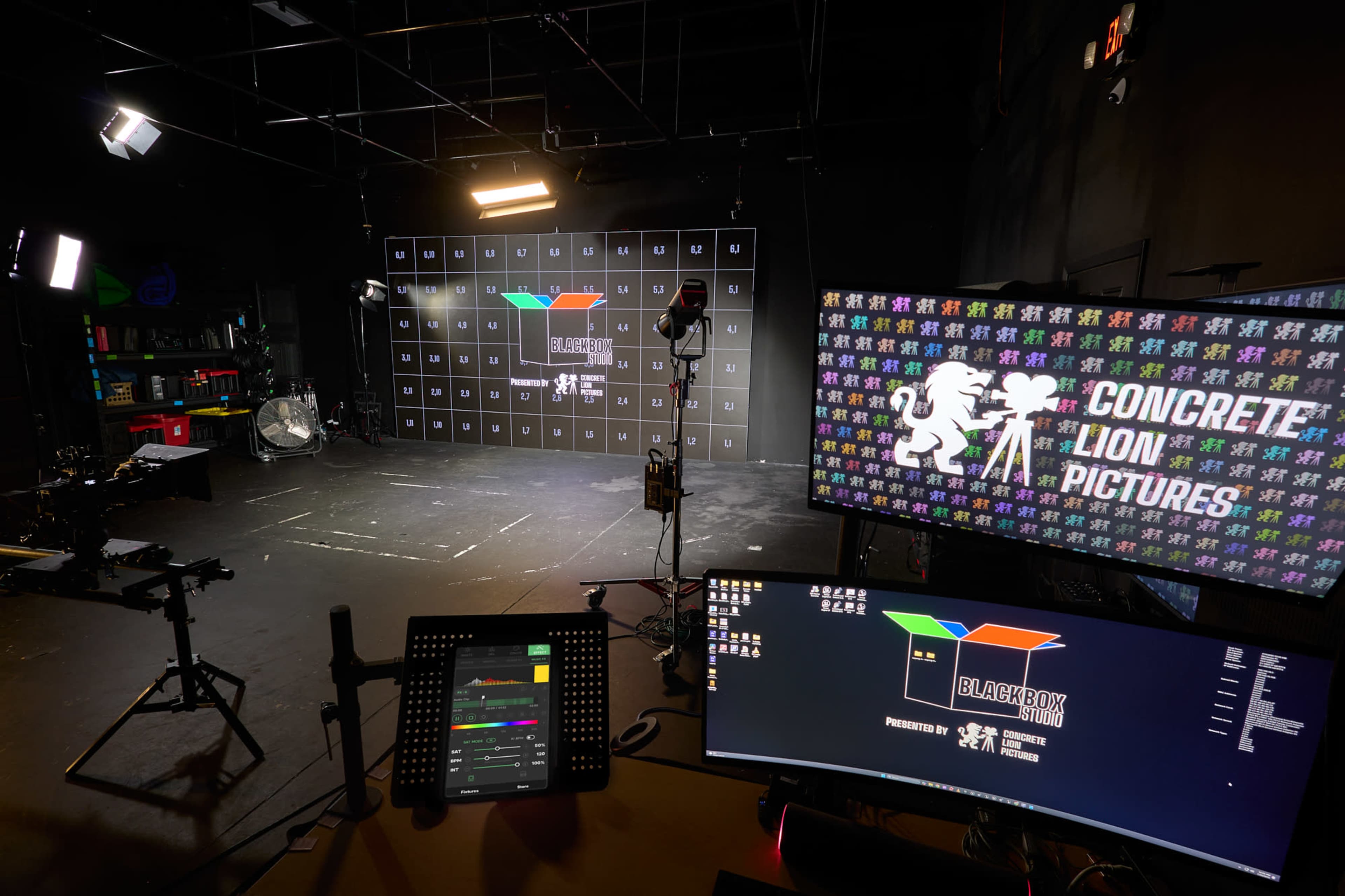 The image shows a studio setting with multiple lighting setups, a large backdrop displaying colorful graphics, and monitoring equipment on a desk.