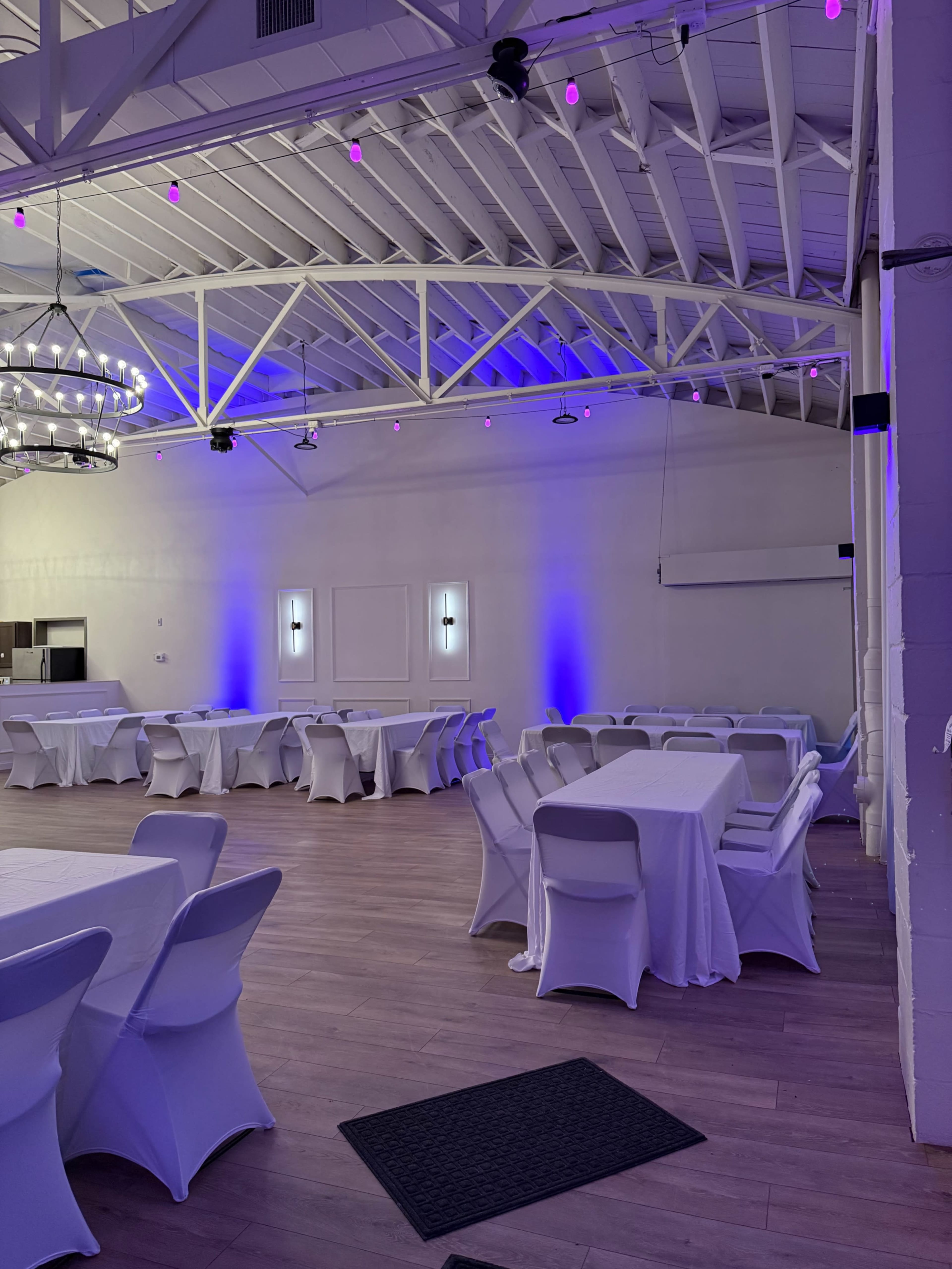 The image shows a spacious event venue with white tables covered in cloth and purple lighting illuminating the walls and ceiling.