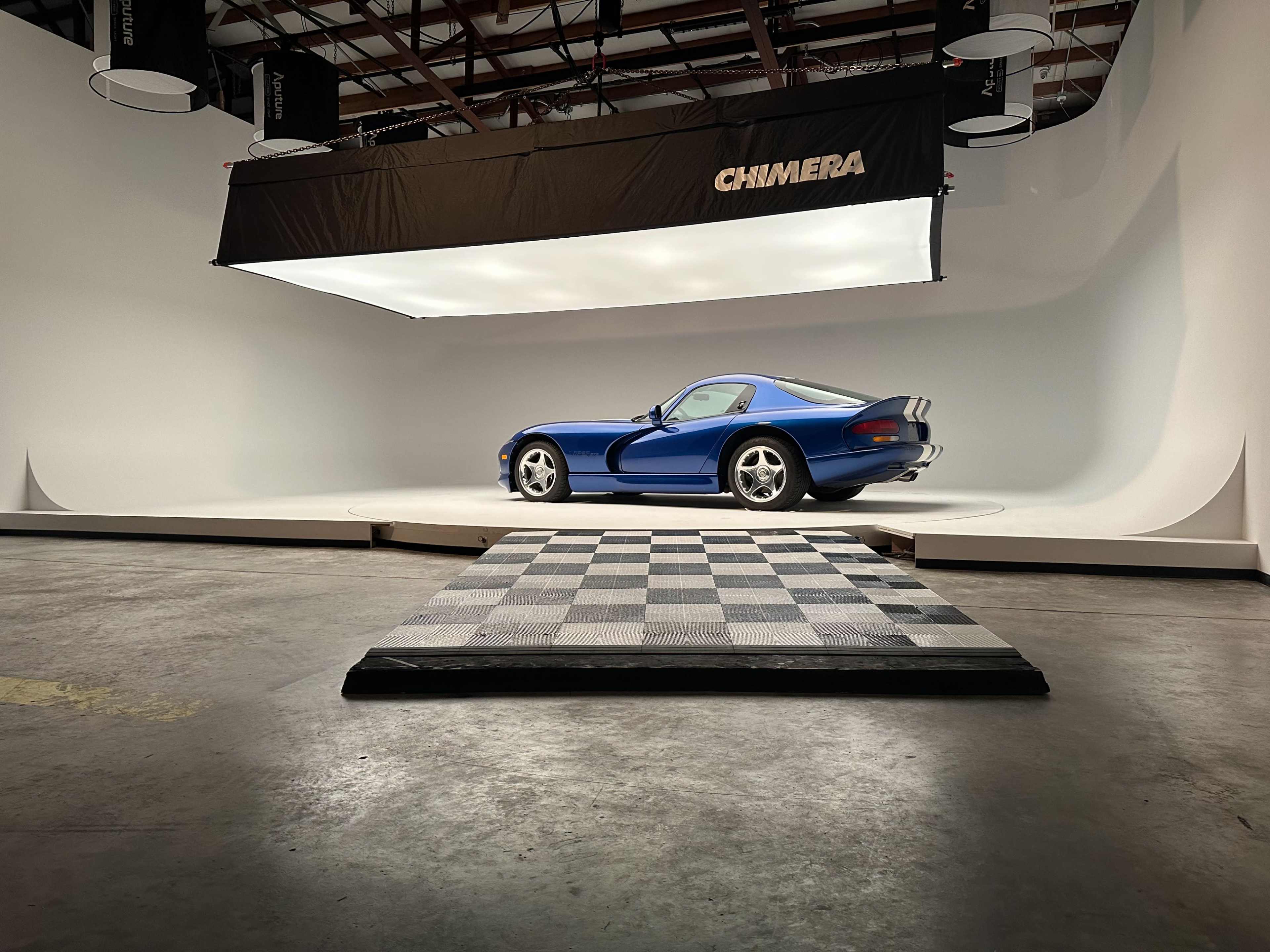 A blue sports car is positioned on a checkered platform in a well-lit photography studio featuring a seamless white backdrop.