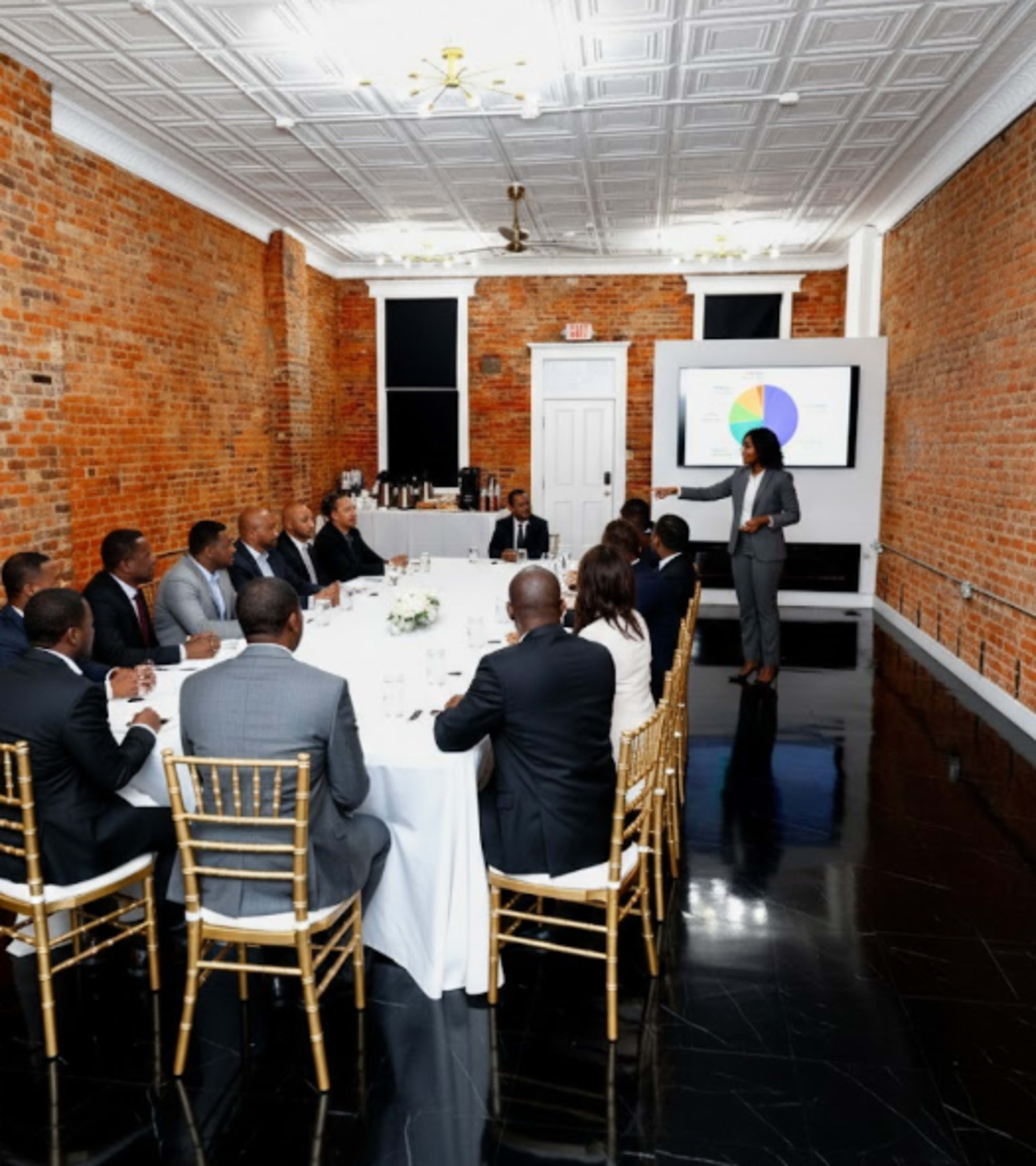 A group of professionals is seated around a rectangular dining table in a room with exposed brick walls, while a speaker presents information using a projector screen.