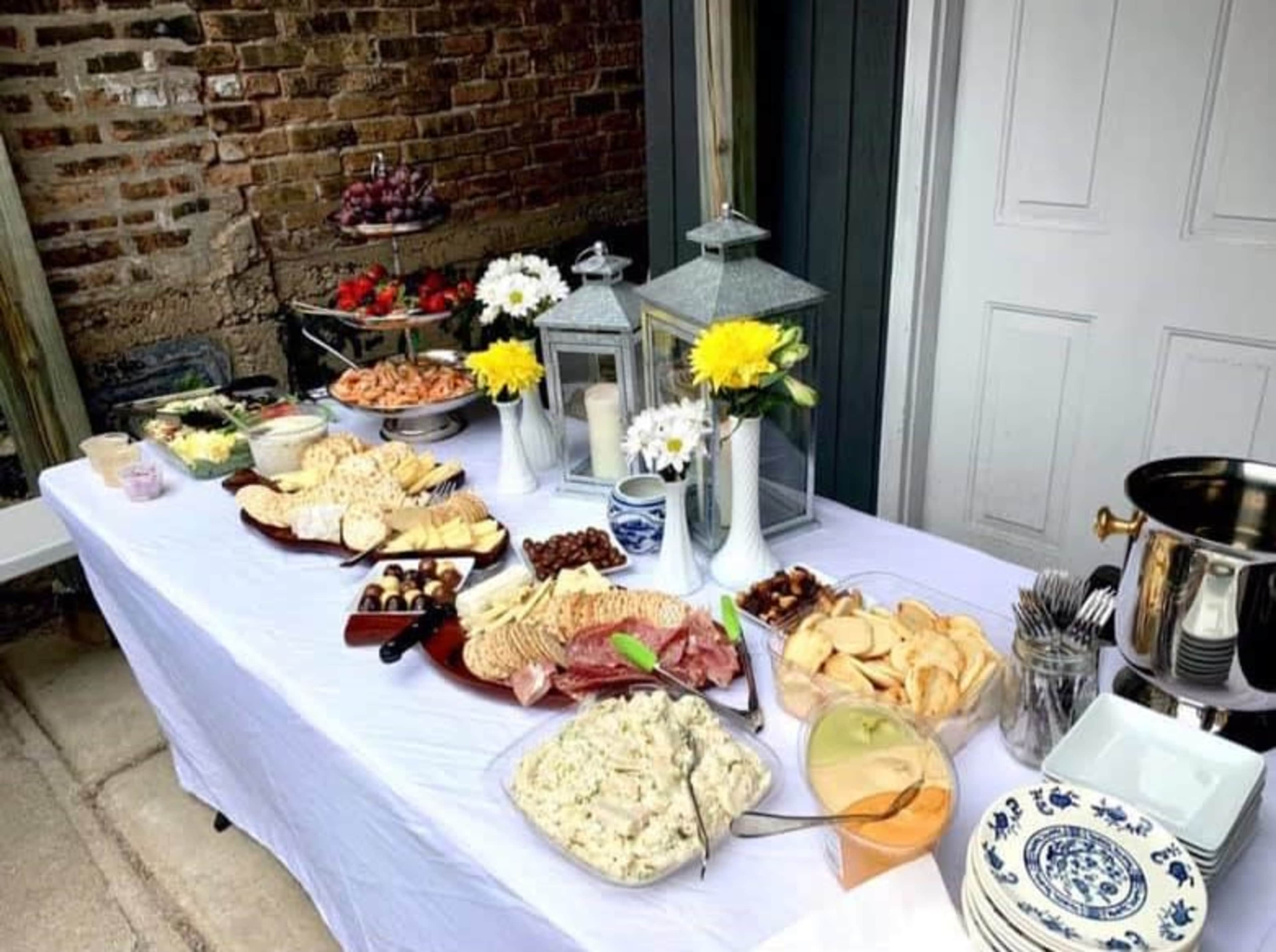 A food buffet is set up on a table featuring various platters of meats, cheeses, vegetables, and salads, along with drinks and decorative centerpieces.