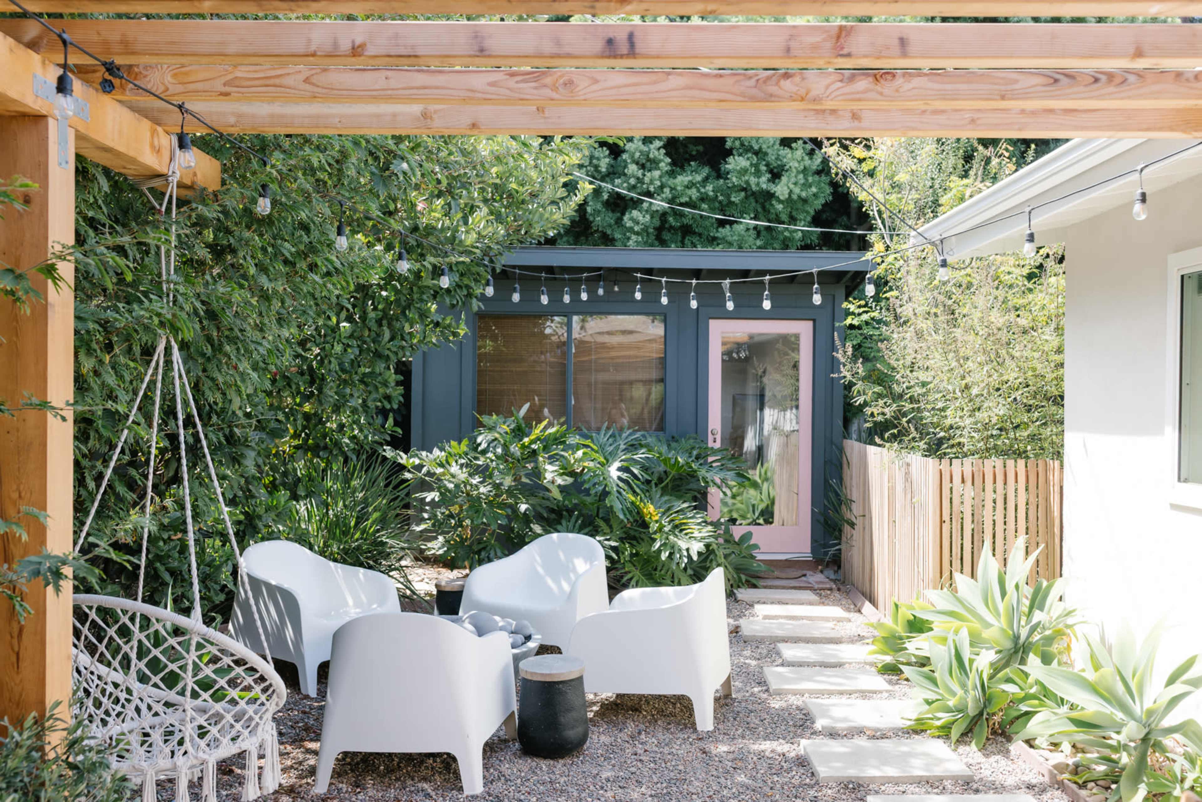 The image features a cozy outdoor seating area with white chairs arranged around a small table, surrounded by greenery, and a decorative hanging swing under a wooden pergola.
