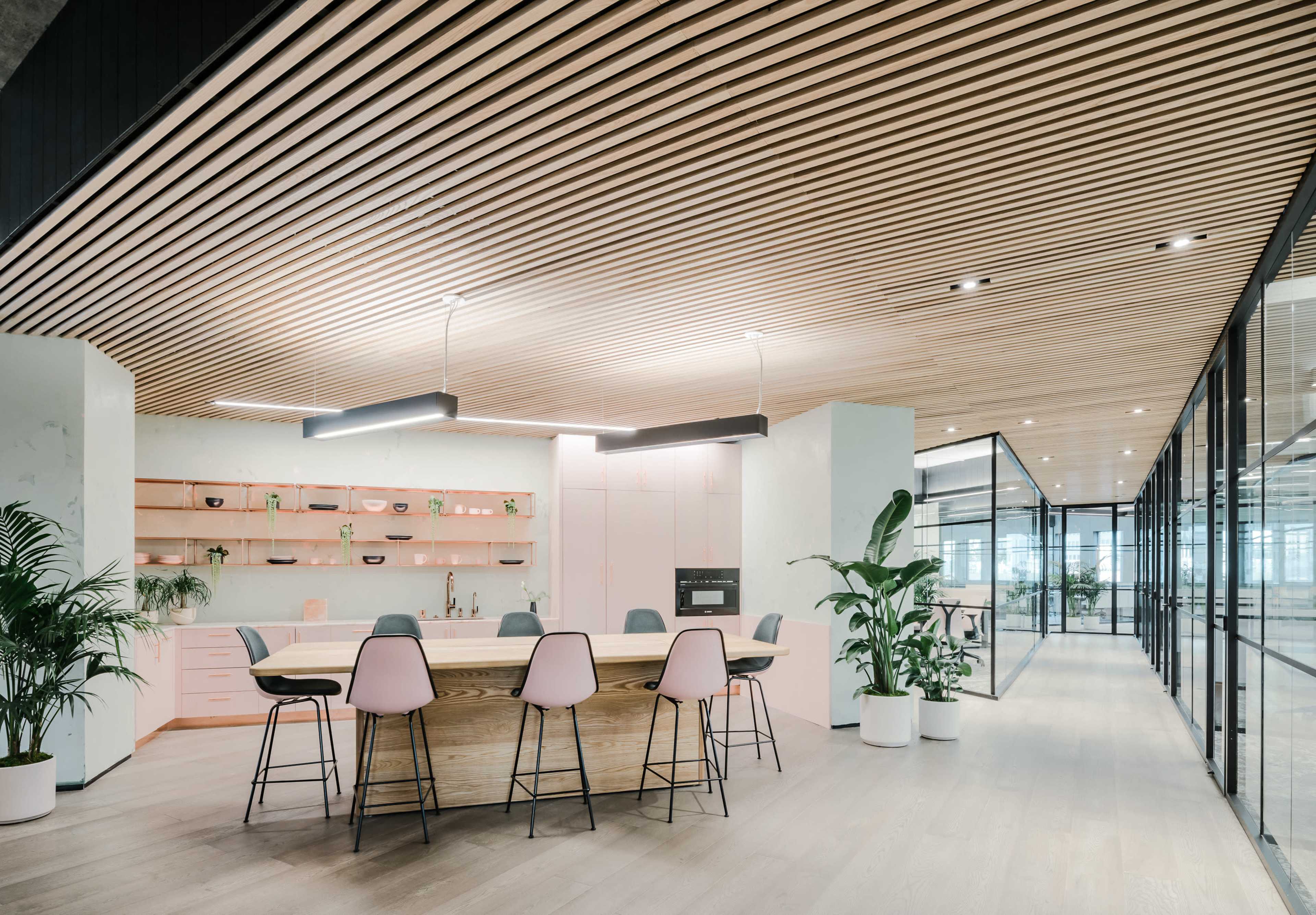 The image shows a modern office kitchen area with a large wooden table surrounded by bar stools, plants, and a contemporary design featuring a light wood ceiling and glass partitions.