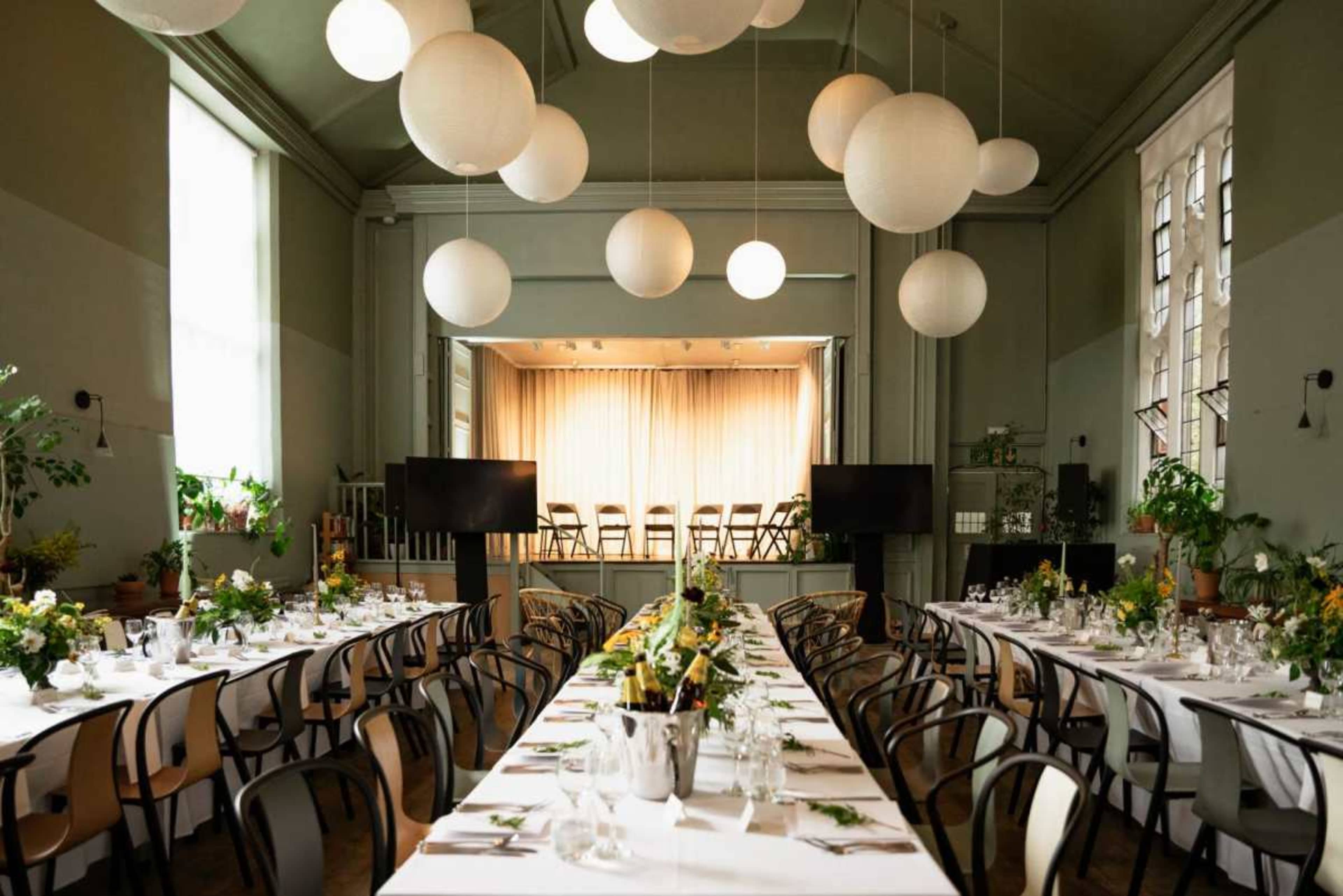 A long banquet table is set for a formal dinner in a room with green walls and large hanging lanterns, facing a small stage with chairs arranged behind a curtain.
