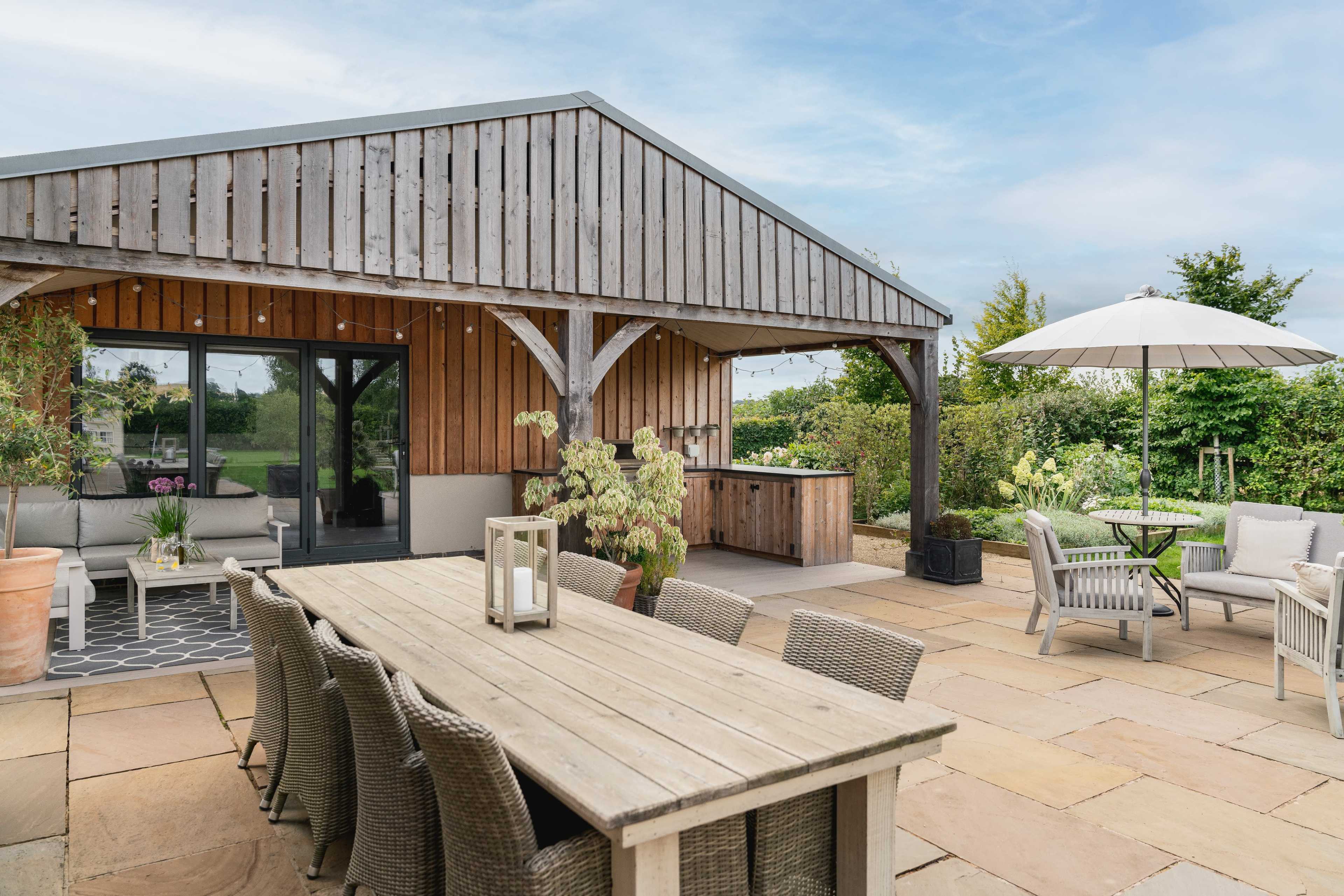 A wooden patio features a large dining table surrounded by chairs, with an umbrella and outdoor seating area beside a garden.