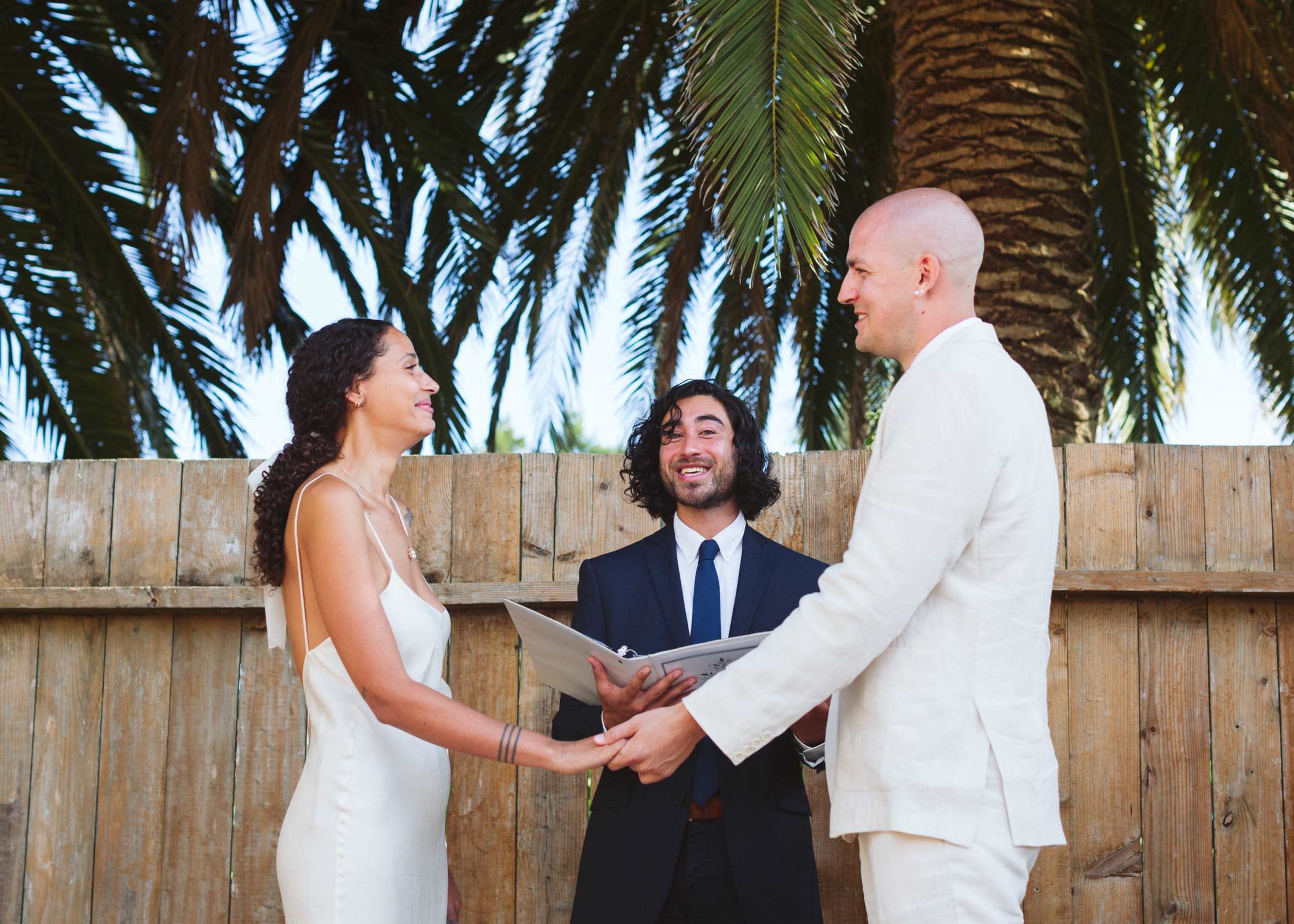 A couple stands in front of a wooden fence under palm trees, exchanging vows while holding hands with a officiant between them.