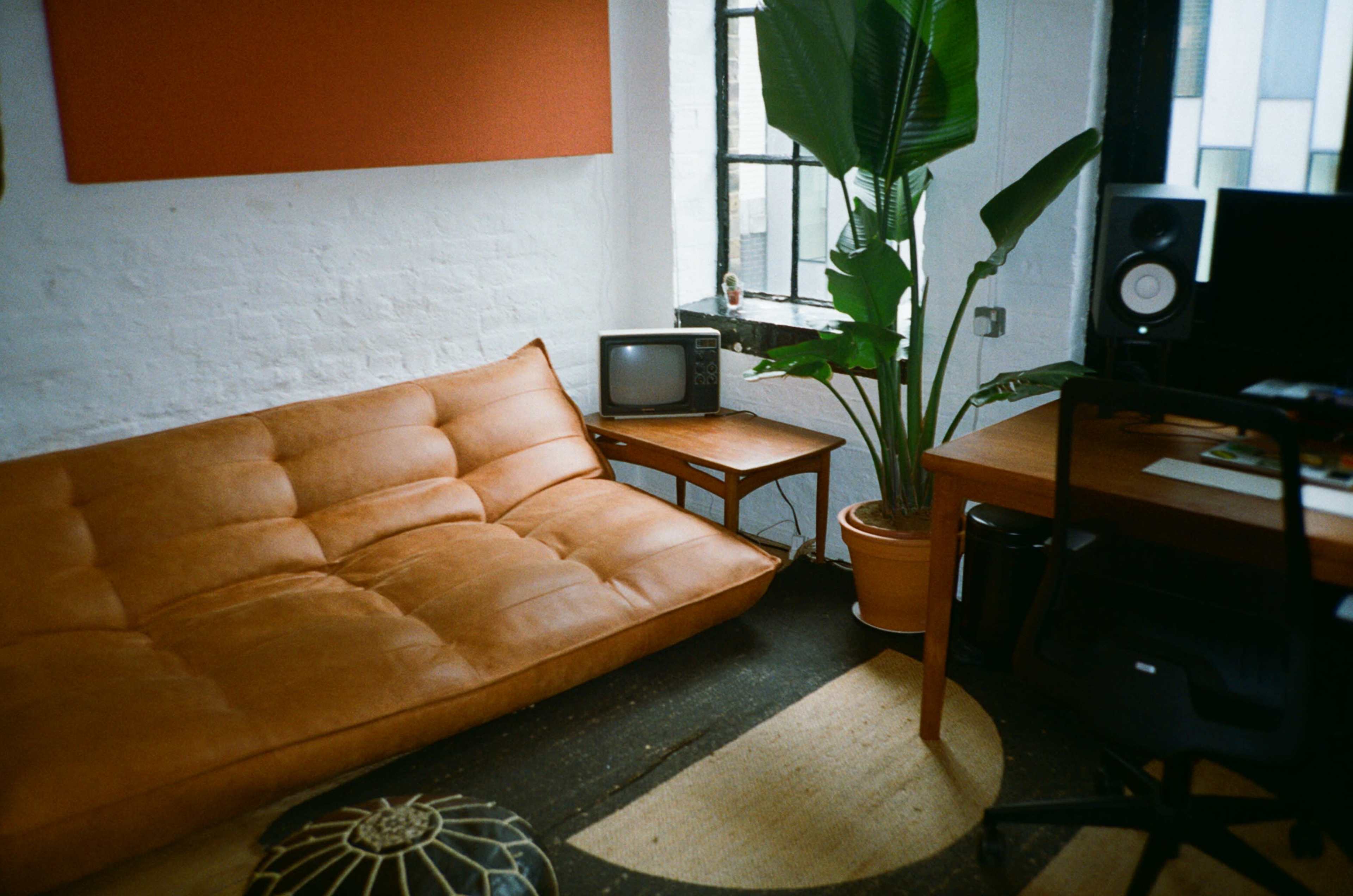A cozy room featuring a brown sofa, a small wooden table, a vintage television, a large potted plant, and a desk with speakers.