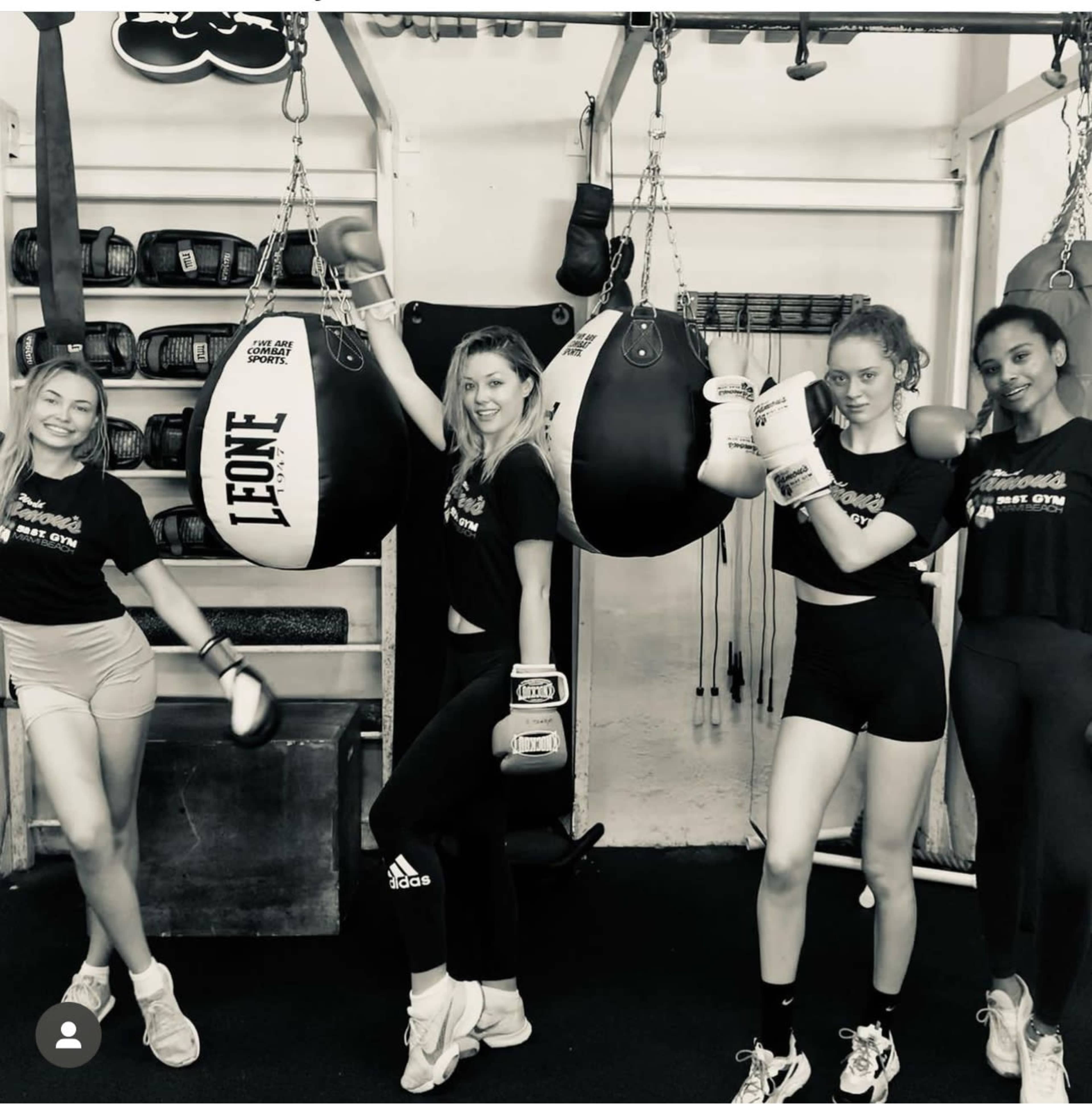 Four women pose together in a boxing gym, each wearing boxing gloves and matching black workout shirts.