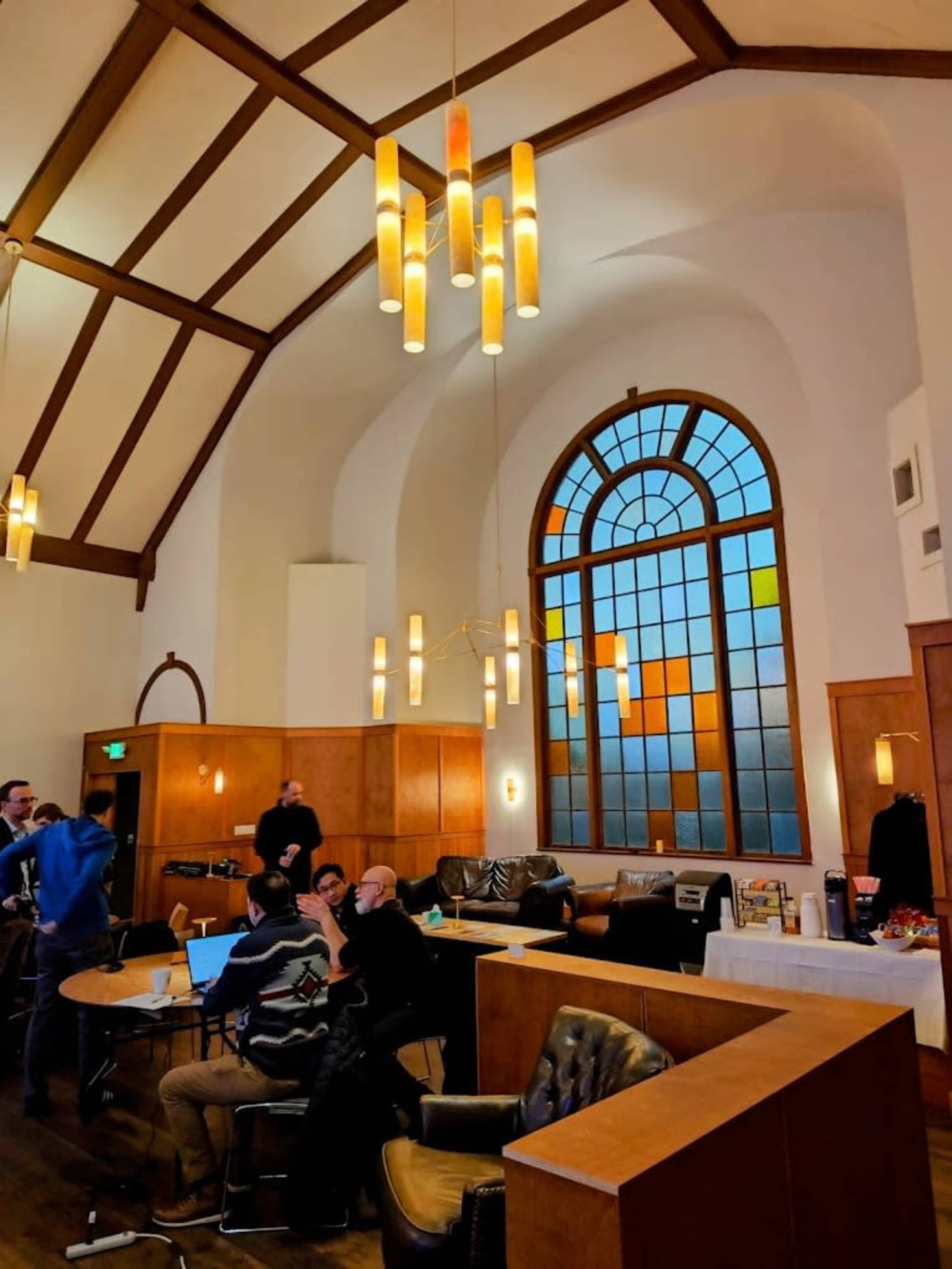 The image shows a spacious interior with high ceilings, featuring large stained glass windows, wooden furniture, and a group of people gathered around tables.
