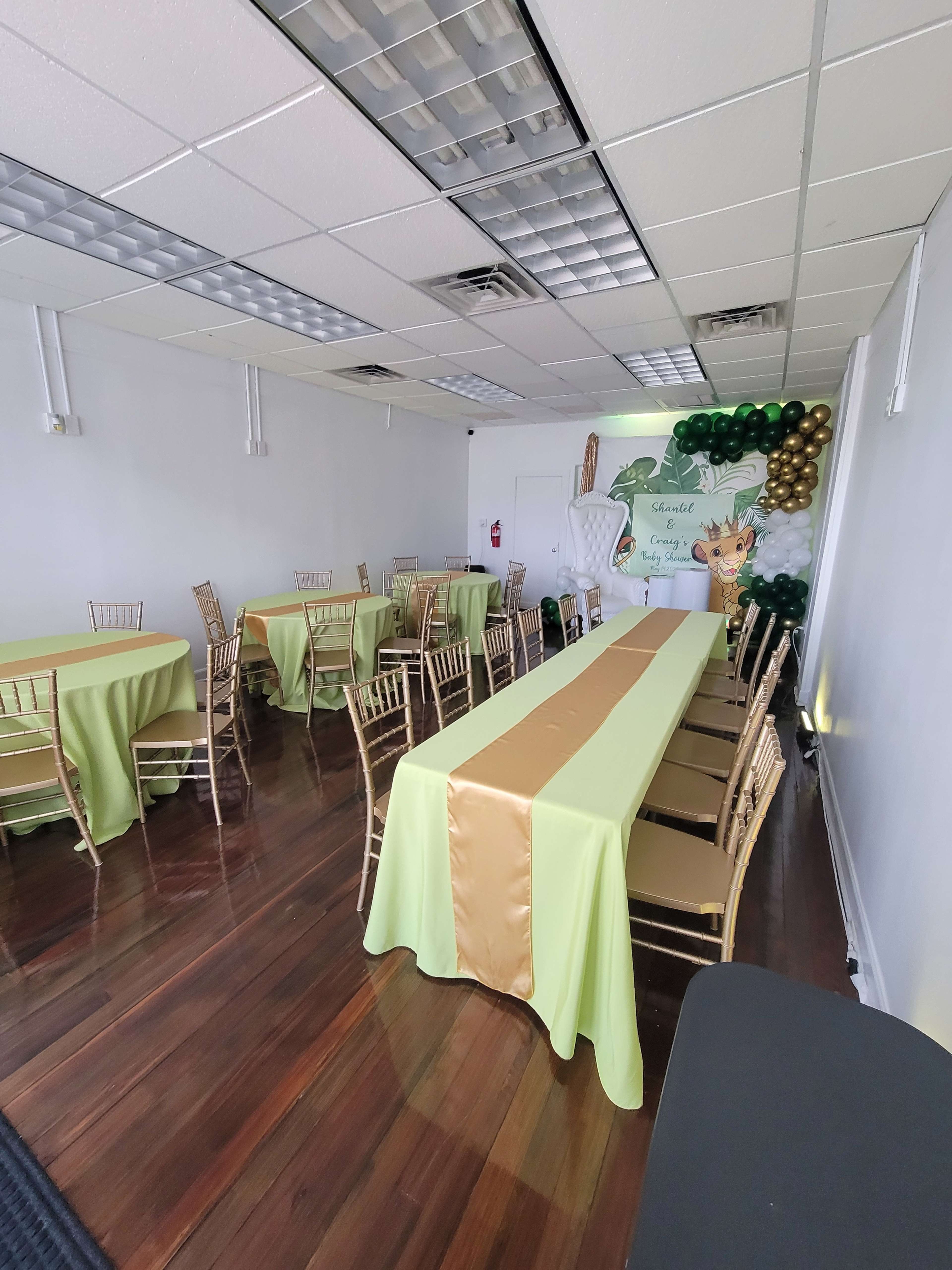 A brightly lit event space features several tables covered in green and gold linens, with decorative balloons and a backdrop on one wall.