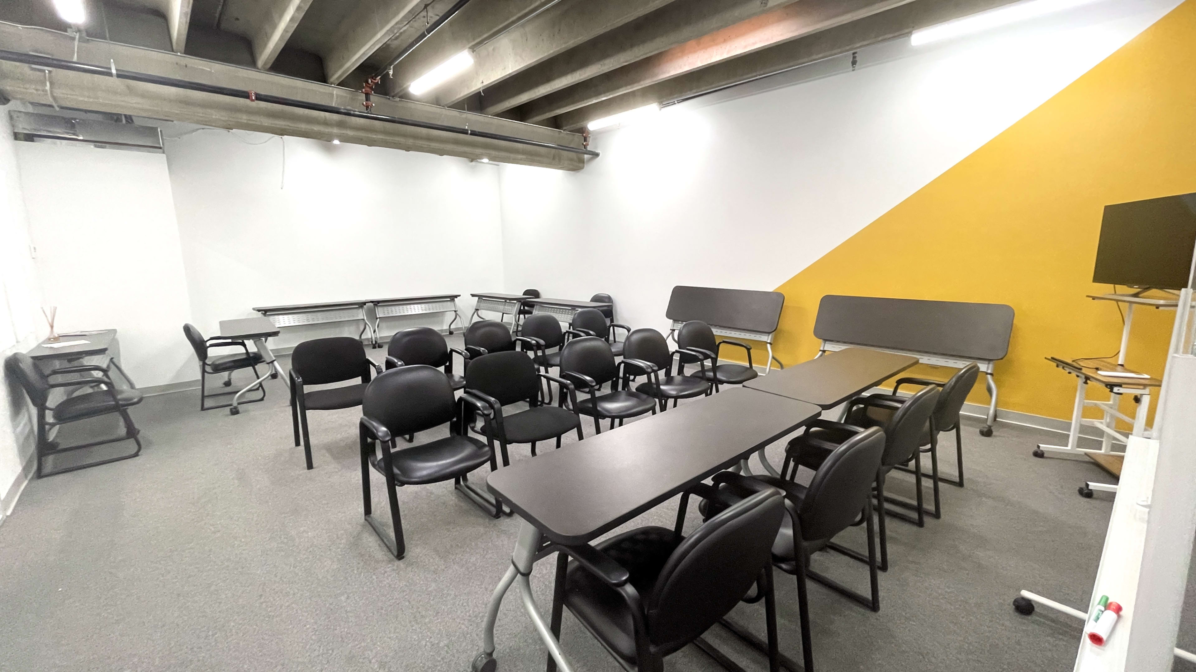 A classroom or meeting space arranged with several black chairs and tables, characterized by a yellow accent wall.