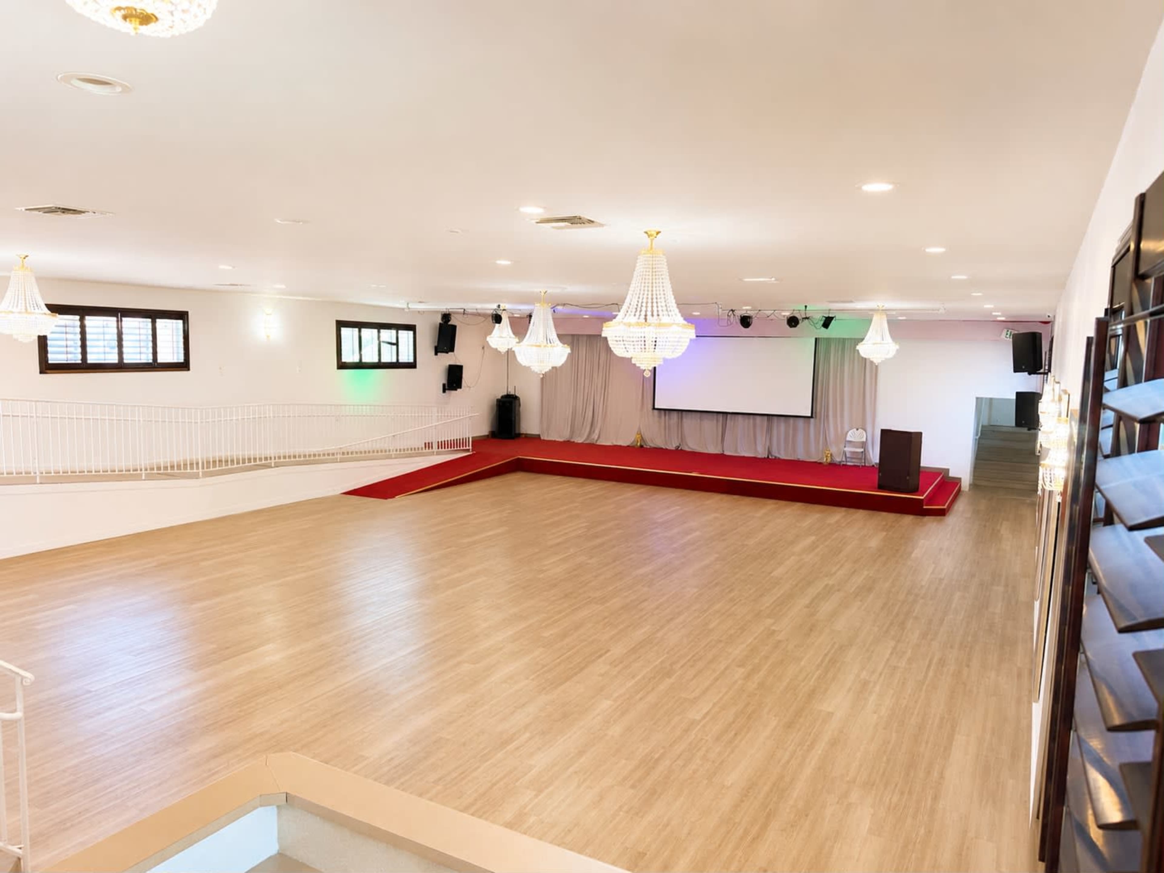 The image shows a spacious event hall with wooden flooring, a raised stage area, and large chandeliers hanging from the ceiling.