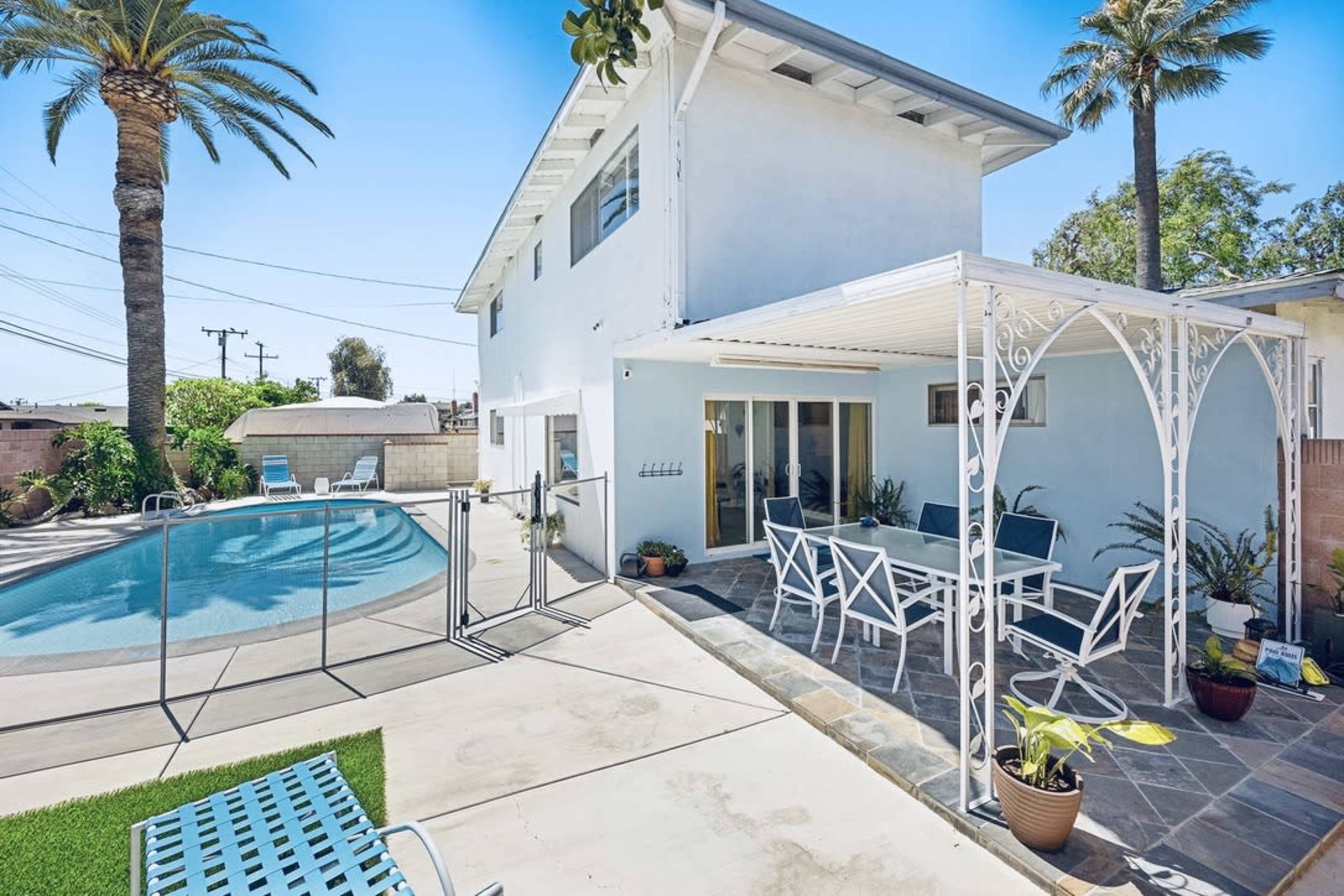A backyard features a swimming pool, a patio with a white metal table and chairs, and palm trees surrounding a two-story blue house.