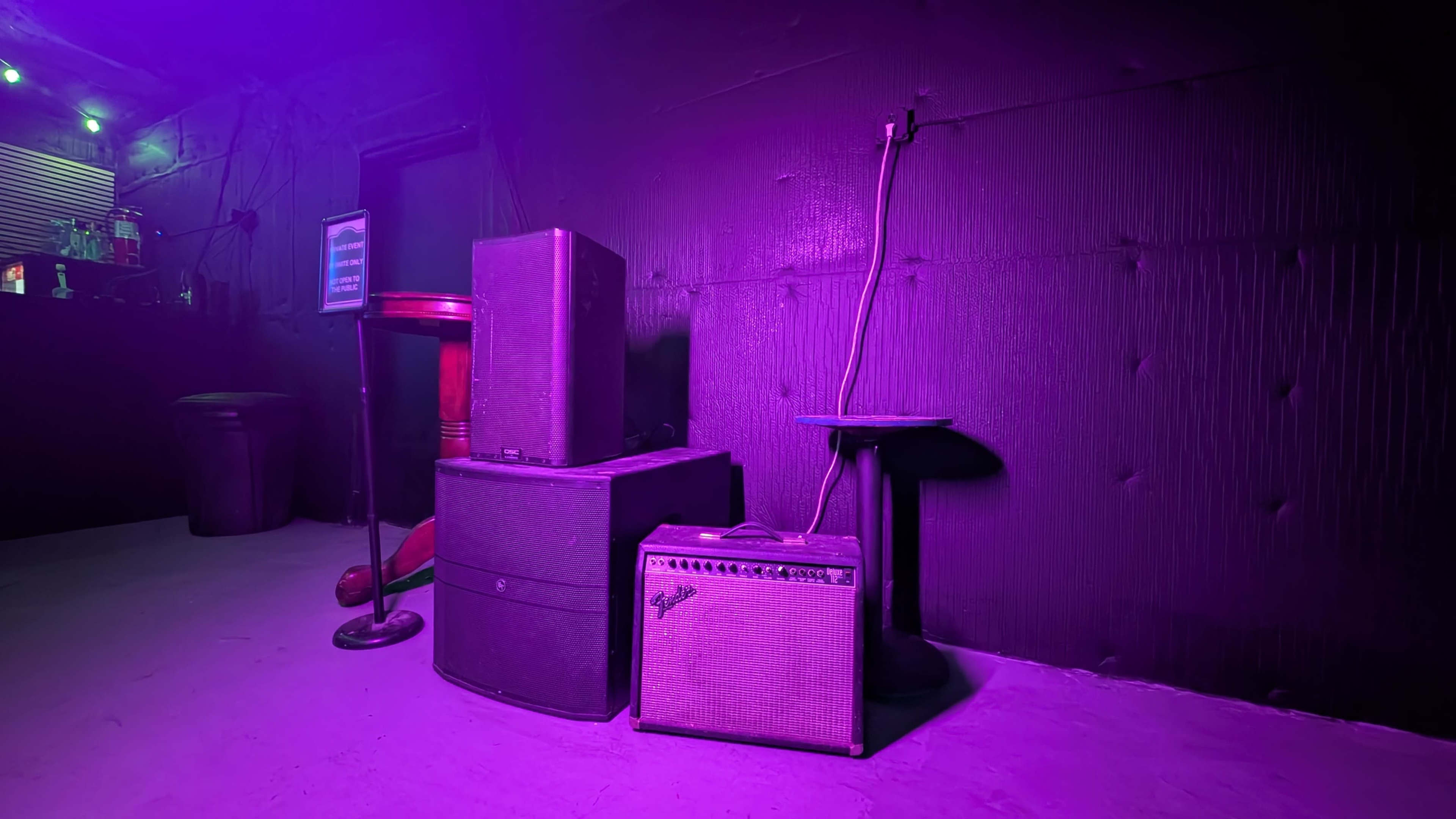The image shows stacked audio equipment, including speakers and an amplifier, against a purple-lit wall in a dimly lit space.