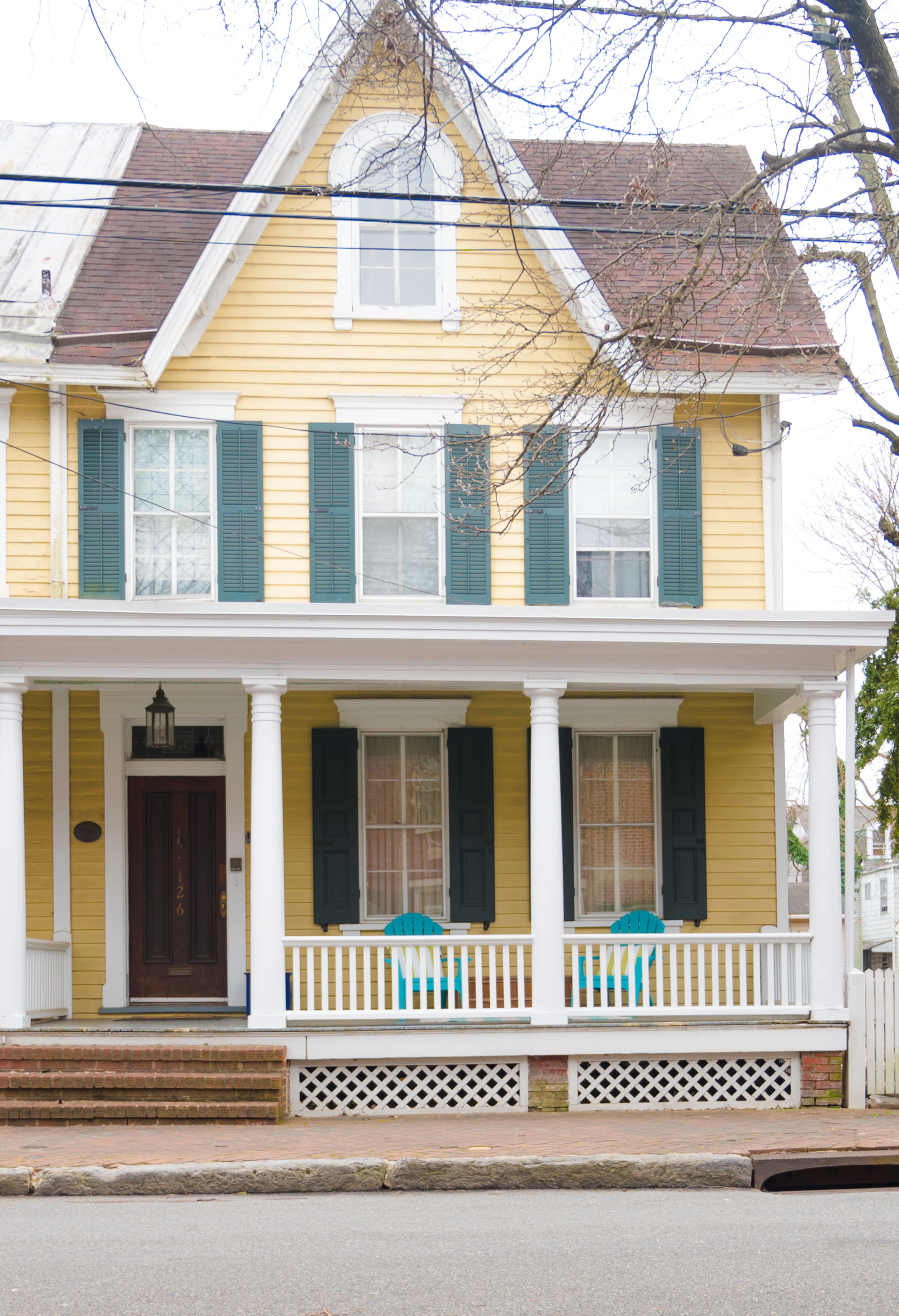 A two-story yellow house with green shutters features a front porch supported by white columns and two blue chairs.