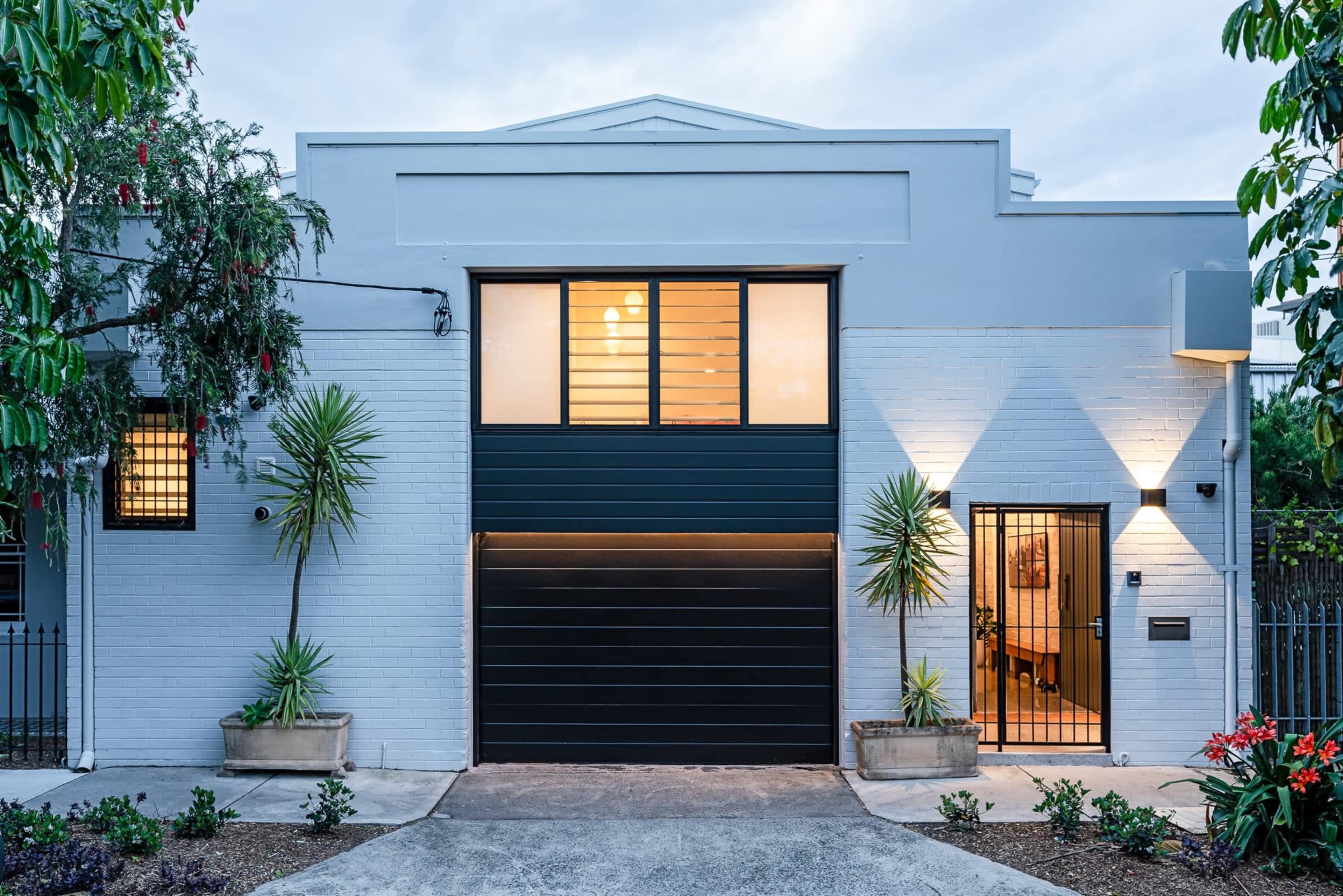 A modern house with a dark garage door, large windows, and tropical plants flanking the entrance.