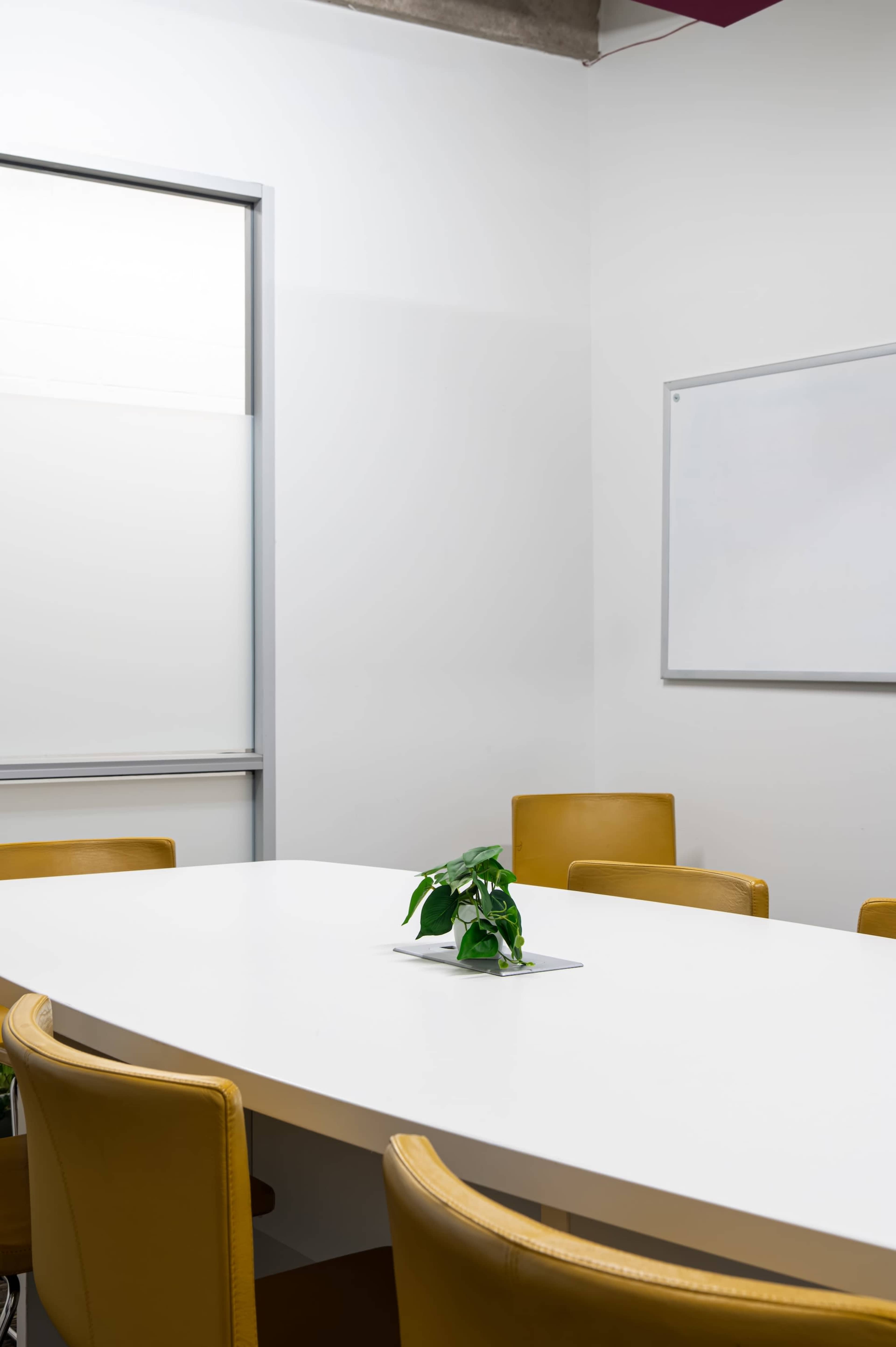 The image shows a minimalistic conference room with a white table, yellow chairs, and a small potted plant in the center, alongside a whiteboard on the wall.