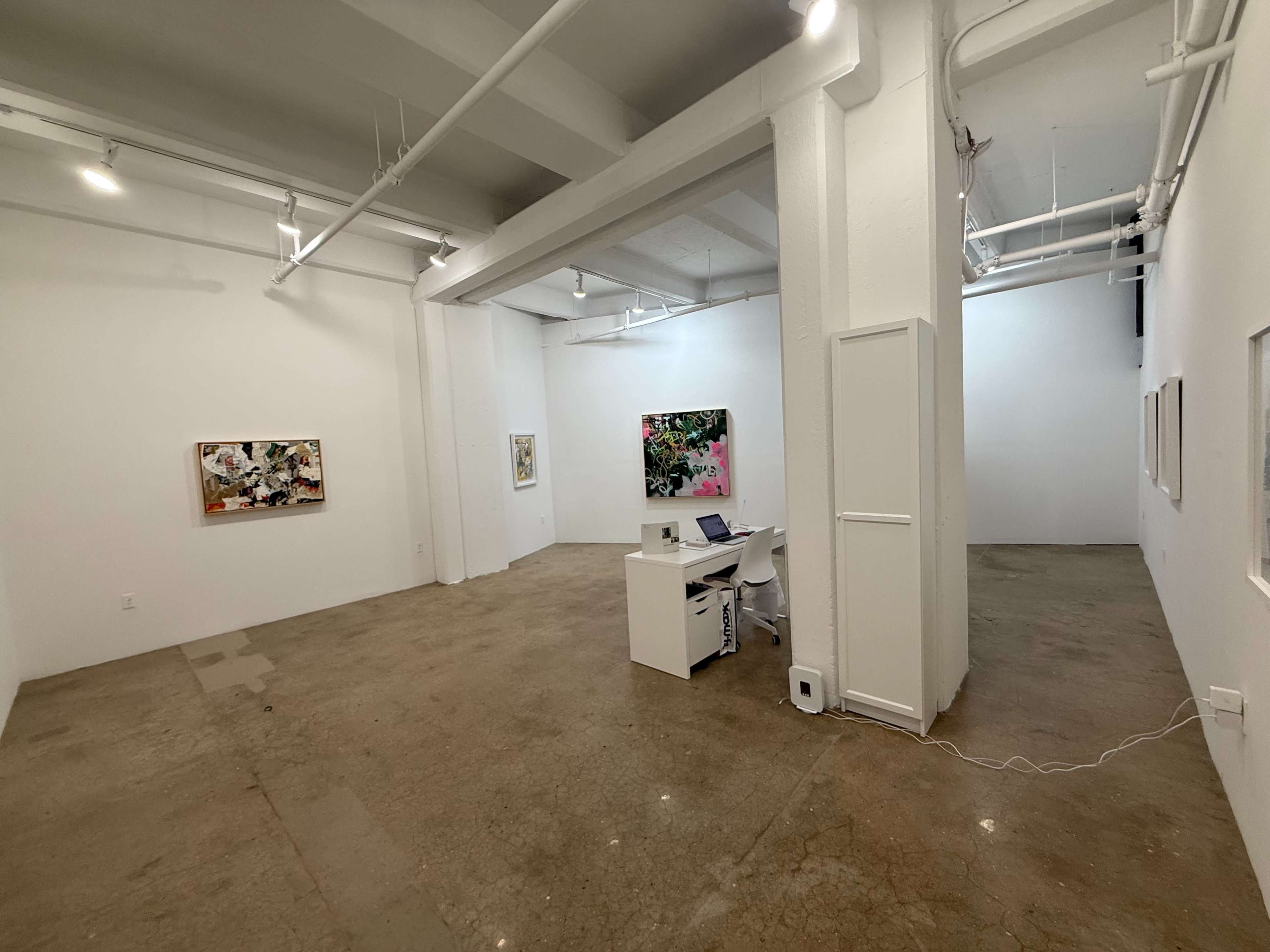 The image shows an art gallery with white walls, concrete flooring, and several pieces of artwork displayed, along with a desk and chair in the foreground.