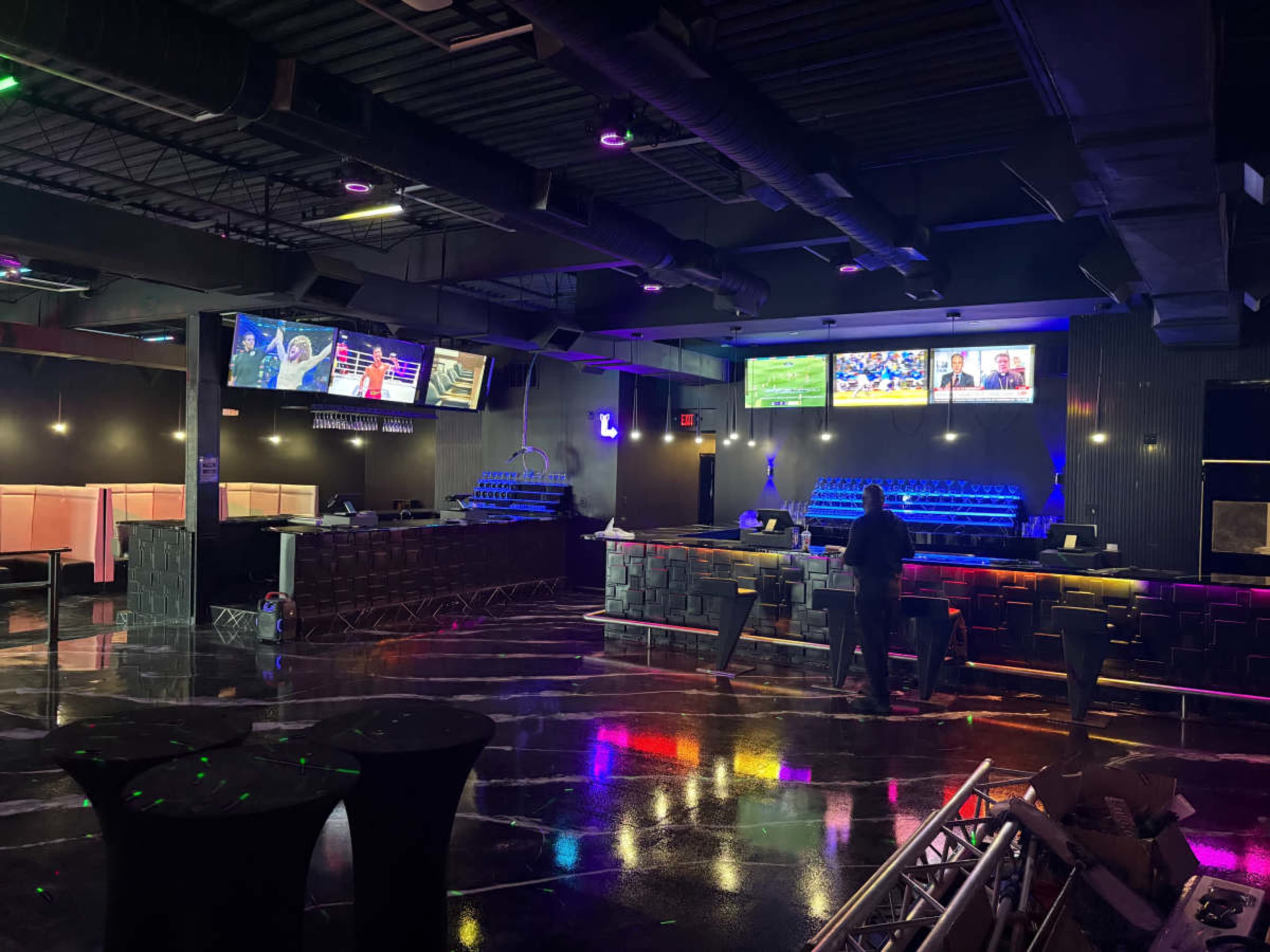The image shows an empty bar area with glossy black floors, multiple televisions displaying sports events, and a long bar counter illuminated by blue lighting.