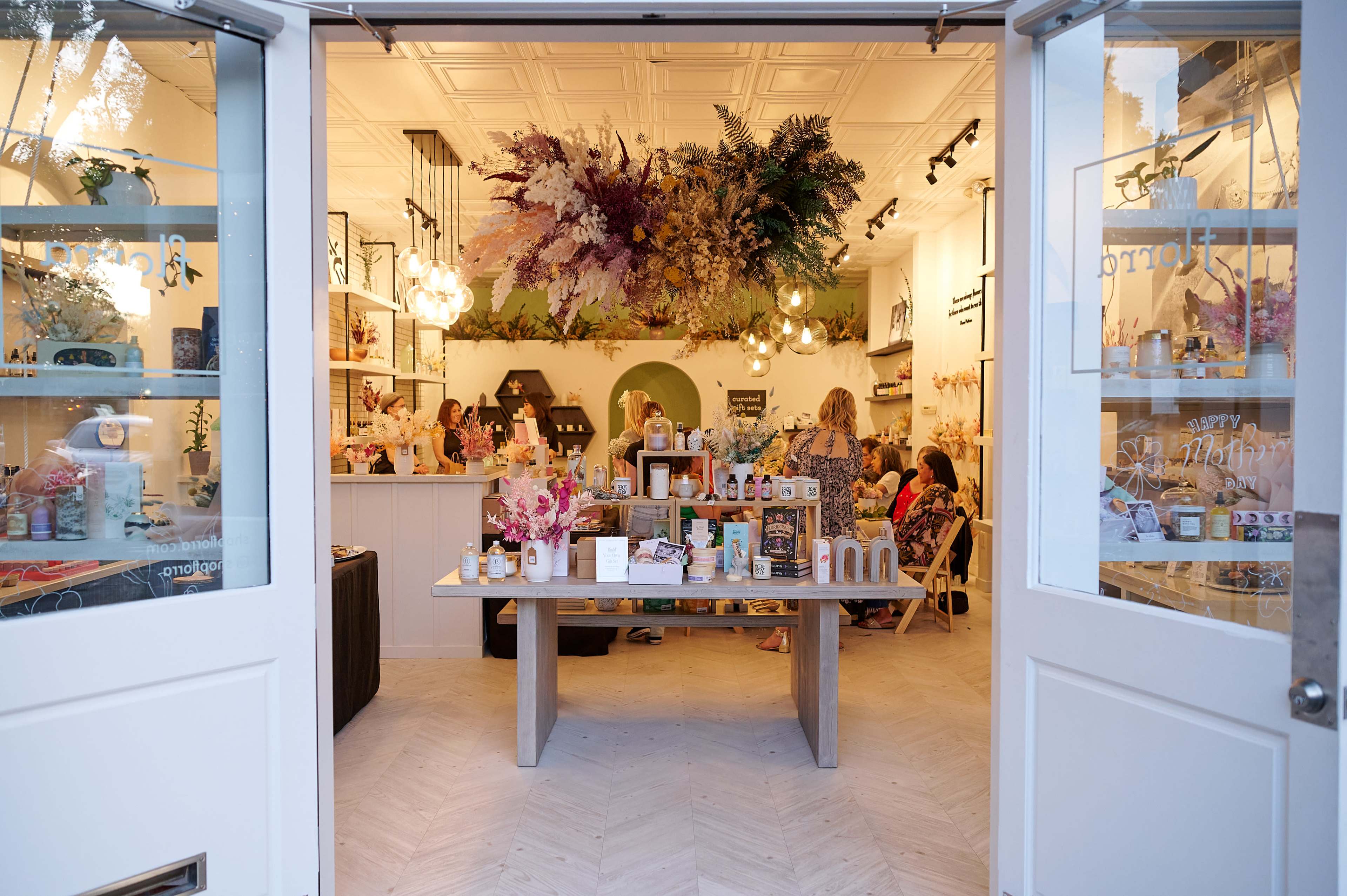 A vibrant store displays various products on a central table, surrounded by customers and adorned with decorative plants hanging from the ceiling.