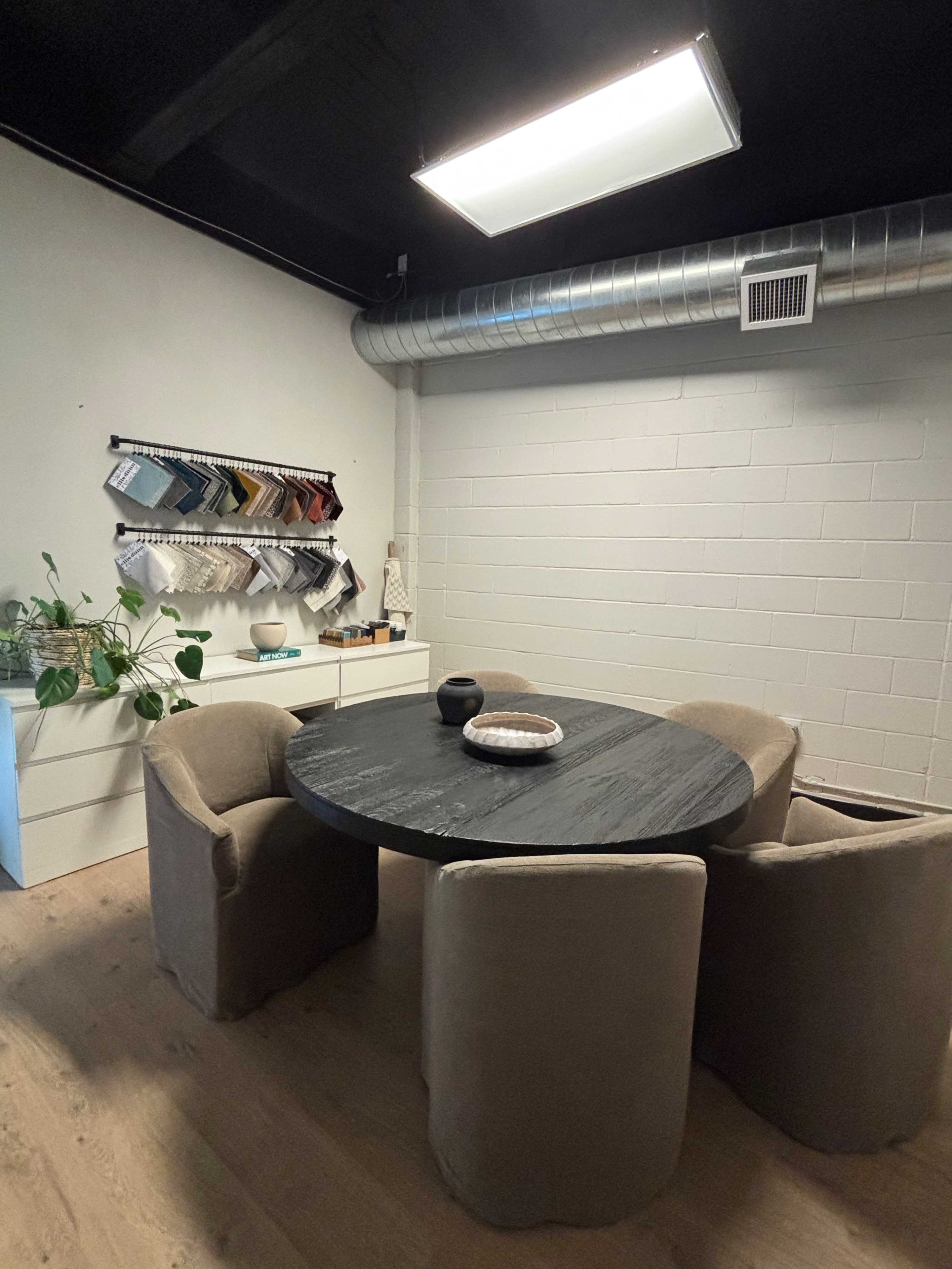 A round black table with four beige upholstered chairs is positioned in a room with a textured wall and a wall-mounted display of fabric samples.