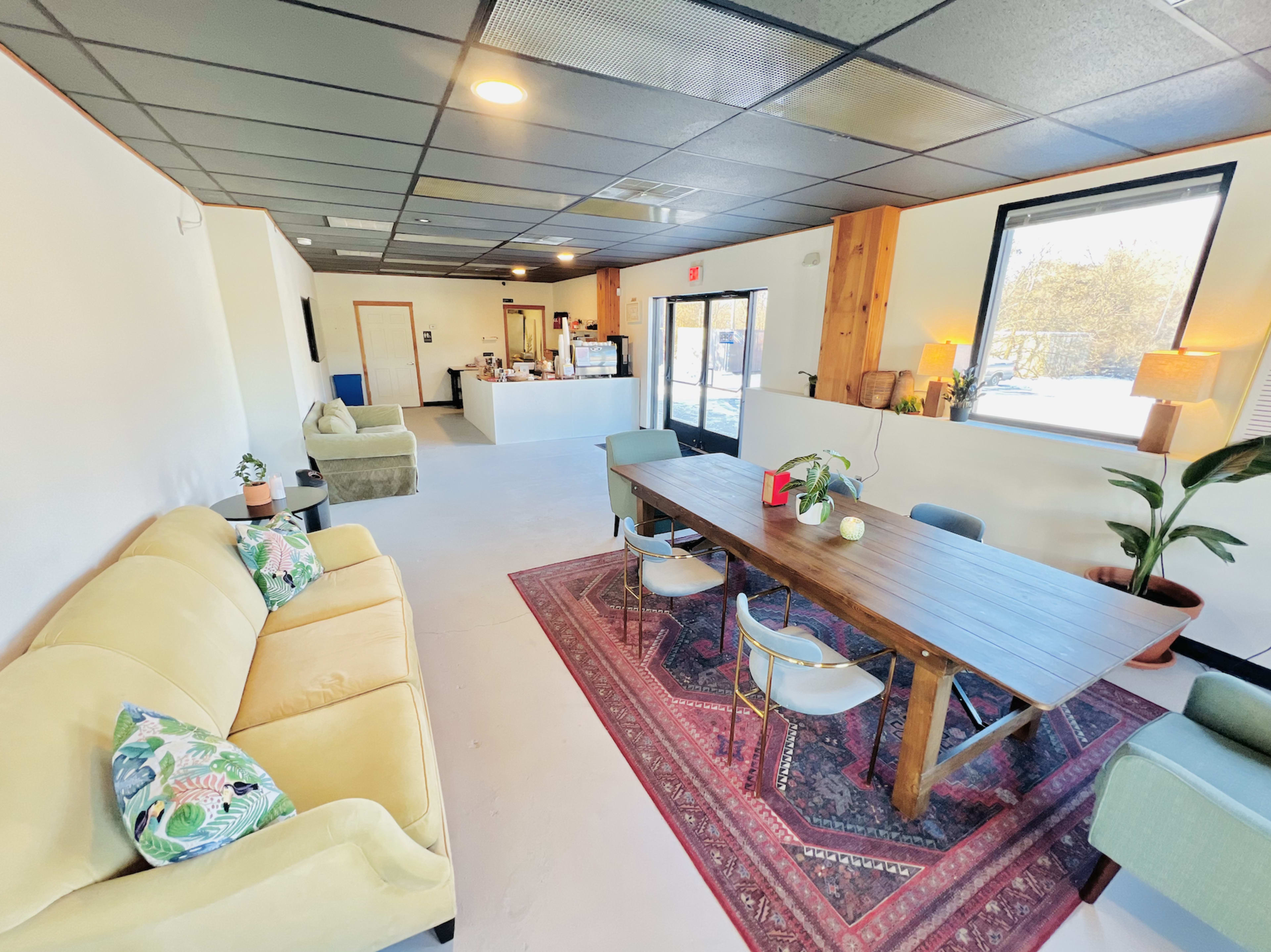 A spacious, brightly lit common area with a large wooden table surrounded by chairs, a sofa, and a kitchenette in the background.