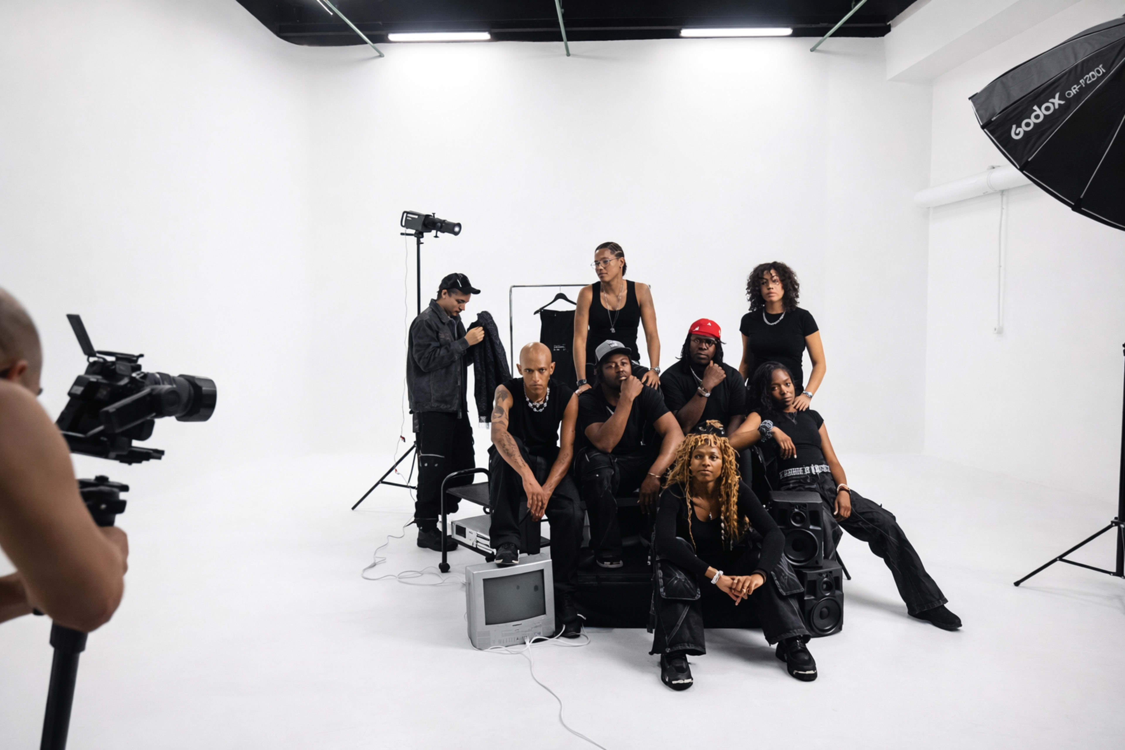 A group of seven individuals poses in a white studio, surrounded by various types of equipment, including cameras and speakers.