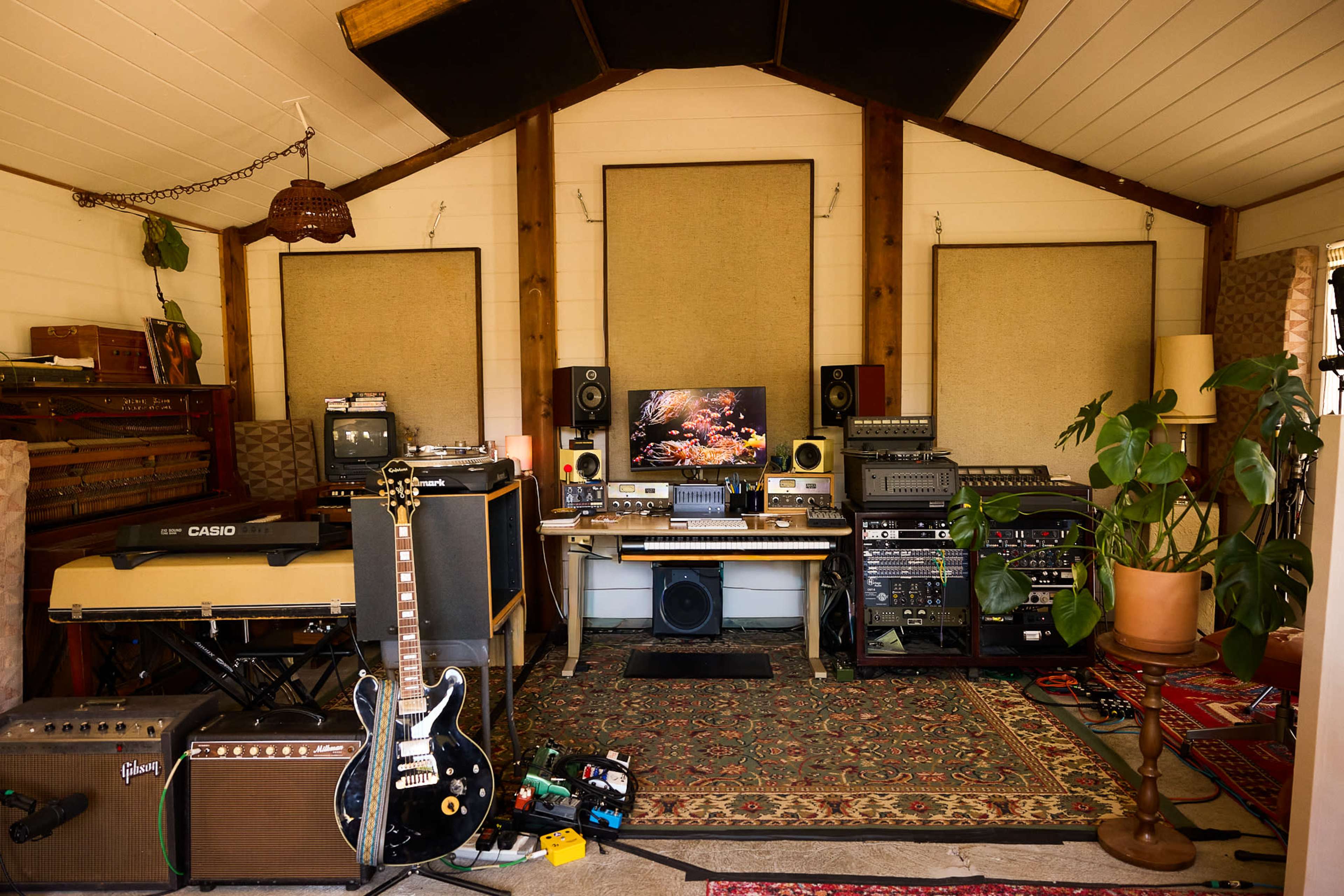 The image shows a cozy music studio space with various audio equipment, including a guitar, speakers, a computer, and a large rug.