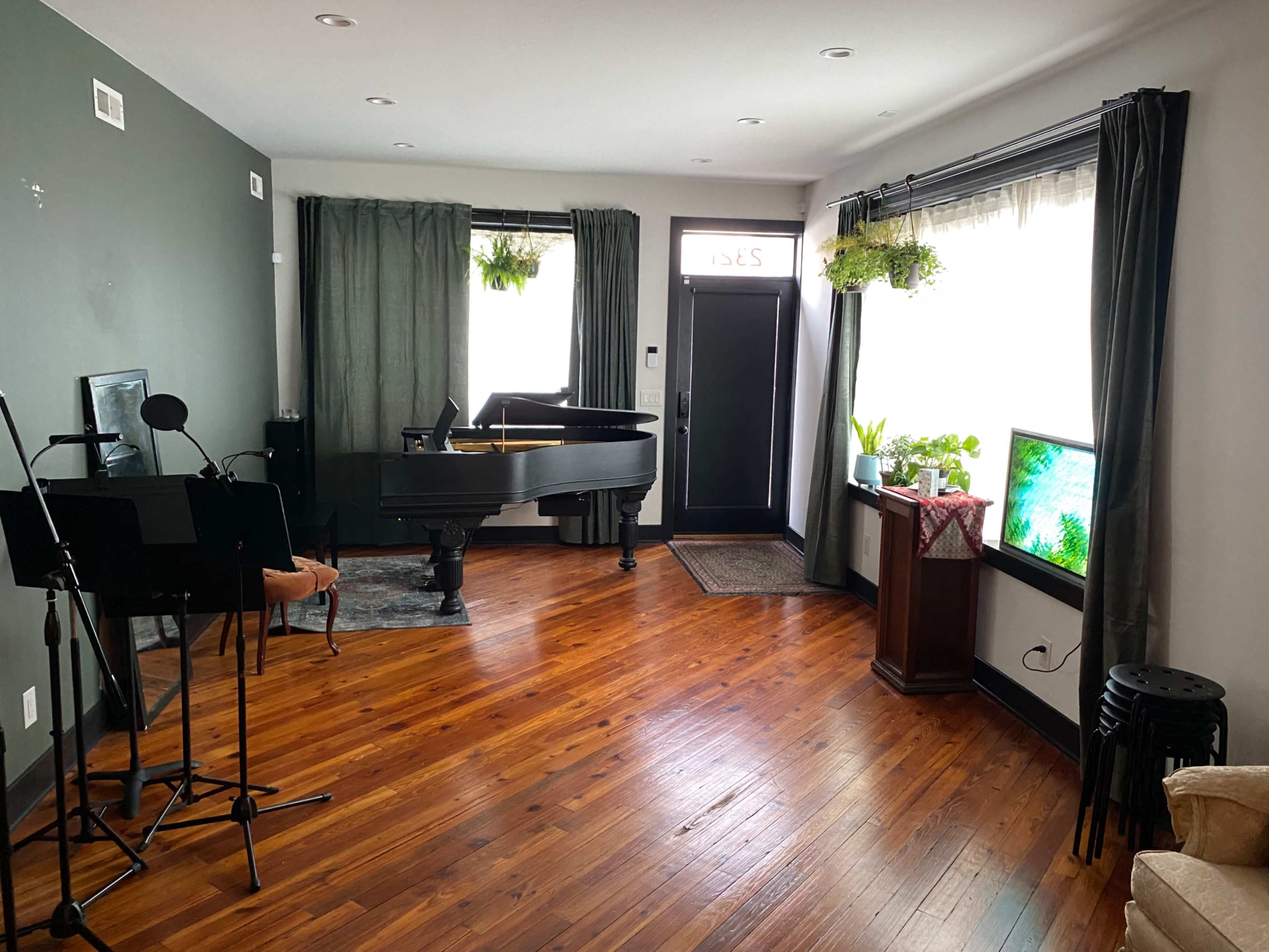 A living room featuring a grand piano, a microphone stand, potted plants, and a television, with hardwood flooring and large windows.